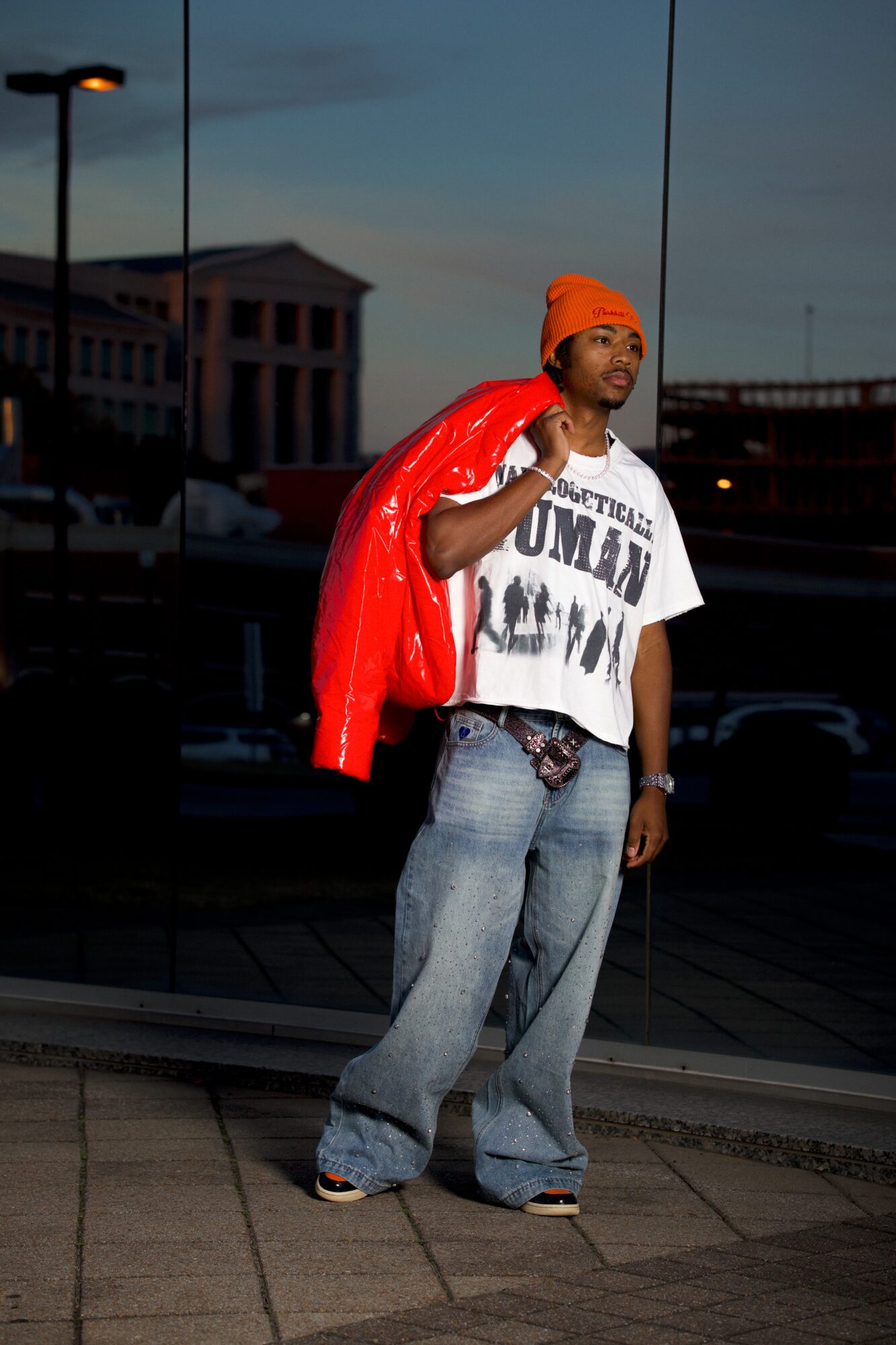 Person standing outdoors at dusk, wearing a white graphic t-shirt, jeans, orange headscarf, and red jacket over shoulder.