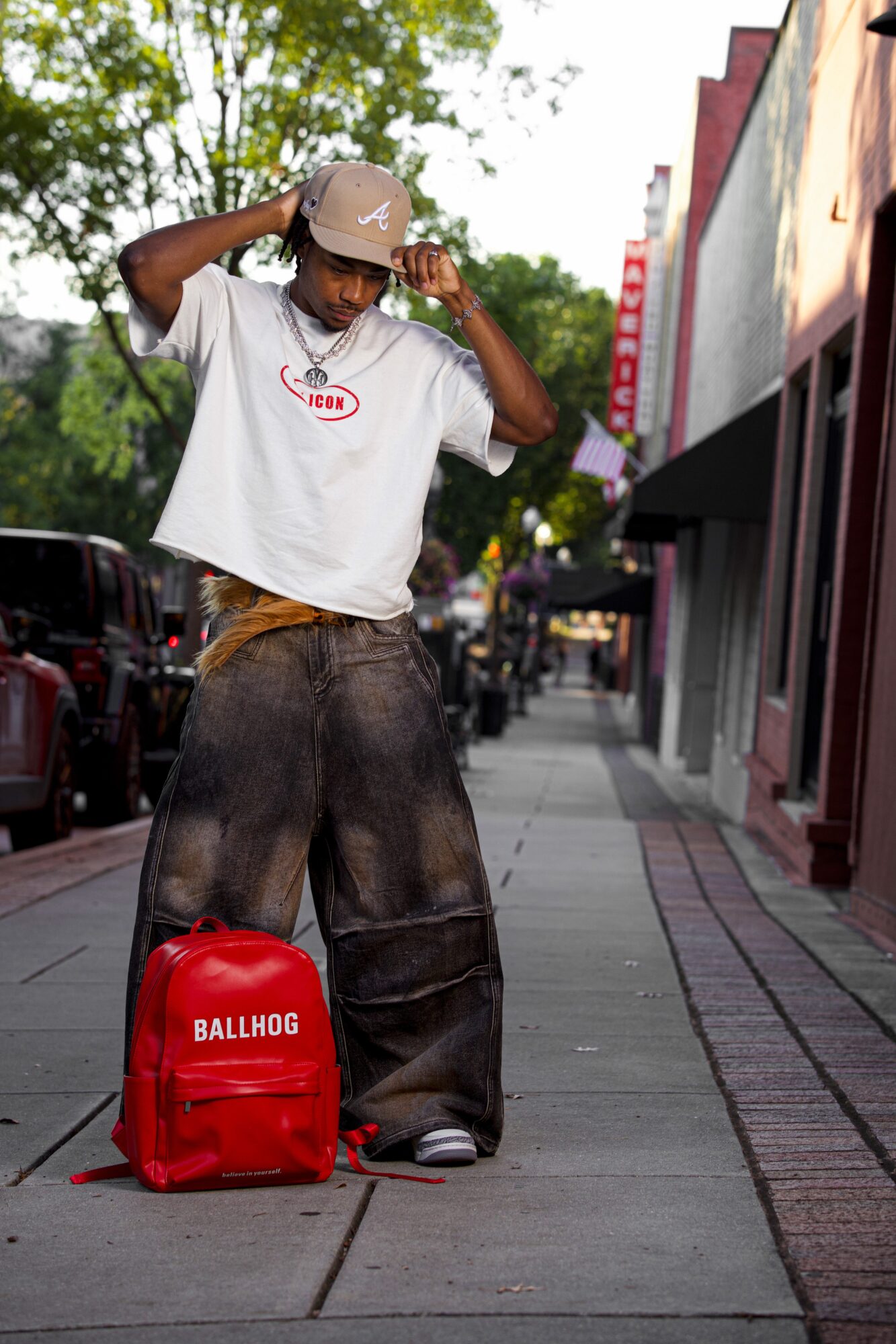 Person standing on sidewalk adjusting hat, wearing oversized pants, white t-shirt, and beige hat, with red backpack nearby.