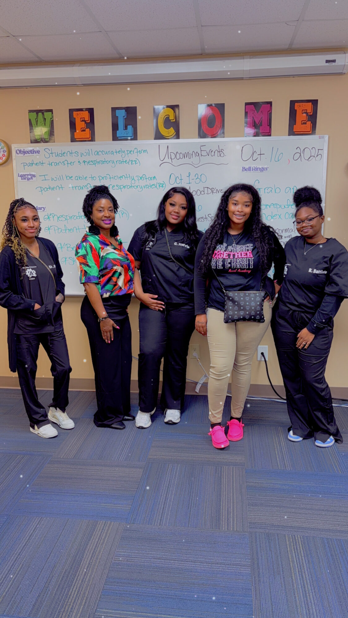 Five young women stand in front of a whiteboard with colorful letters spelling 'WELCOME' and a message, in a classroom.