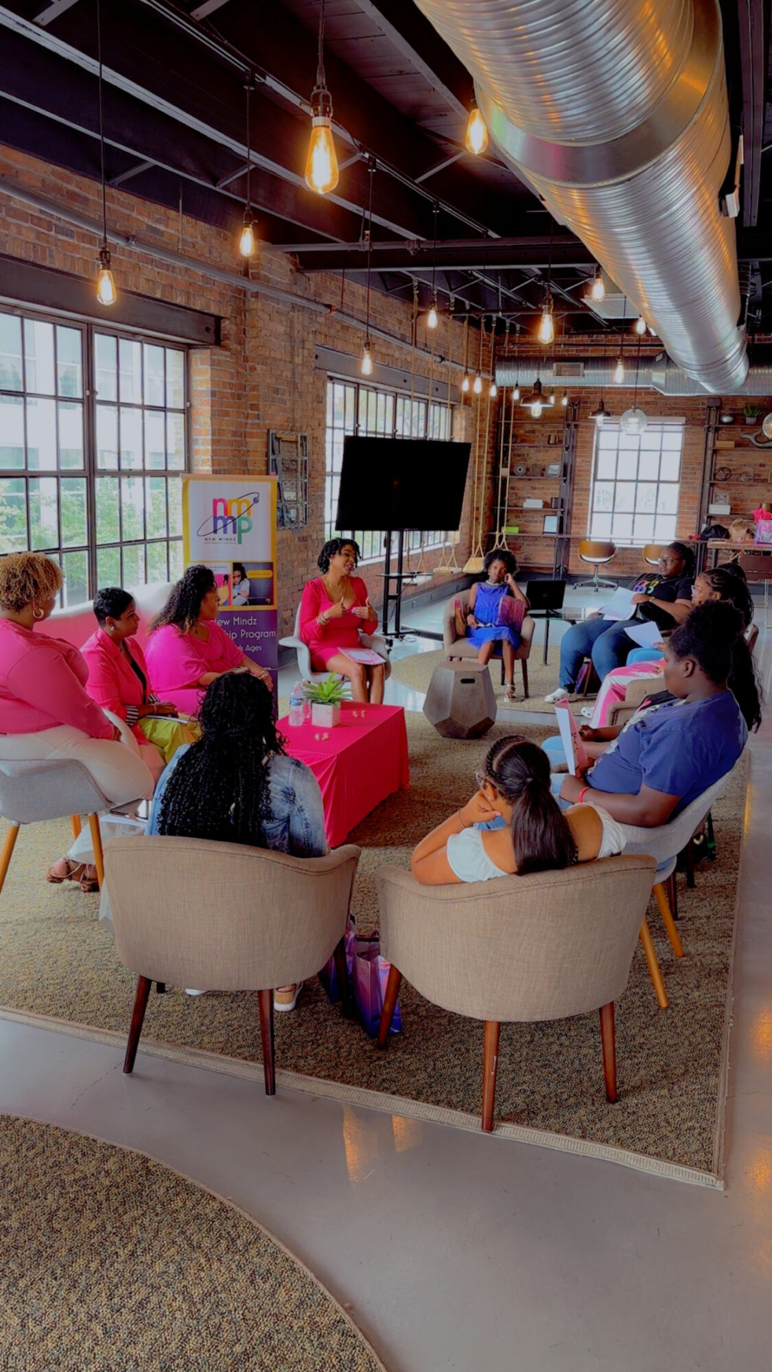 People seated in a circle in a spacious, industrial-style room with large windows and exposed ductwork.
