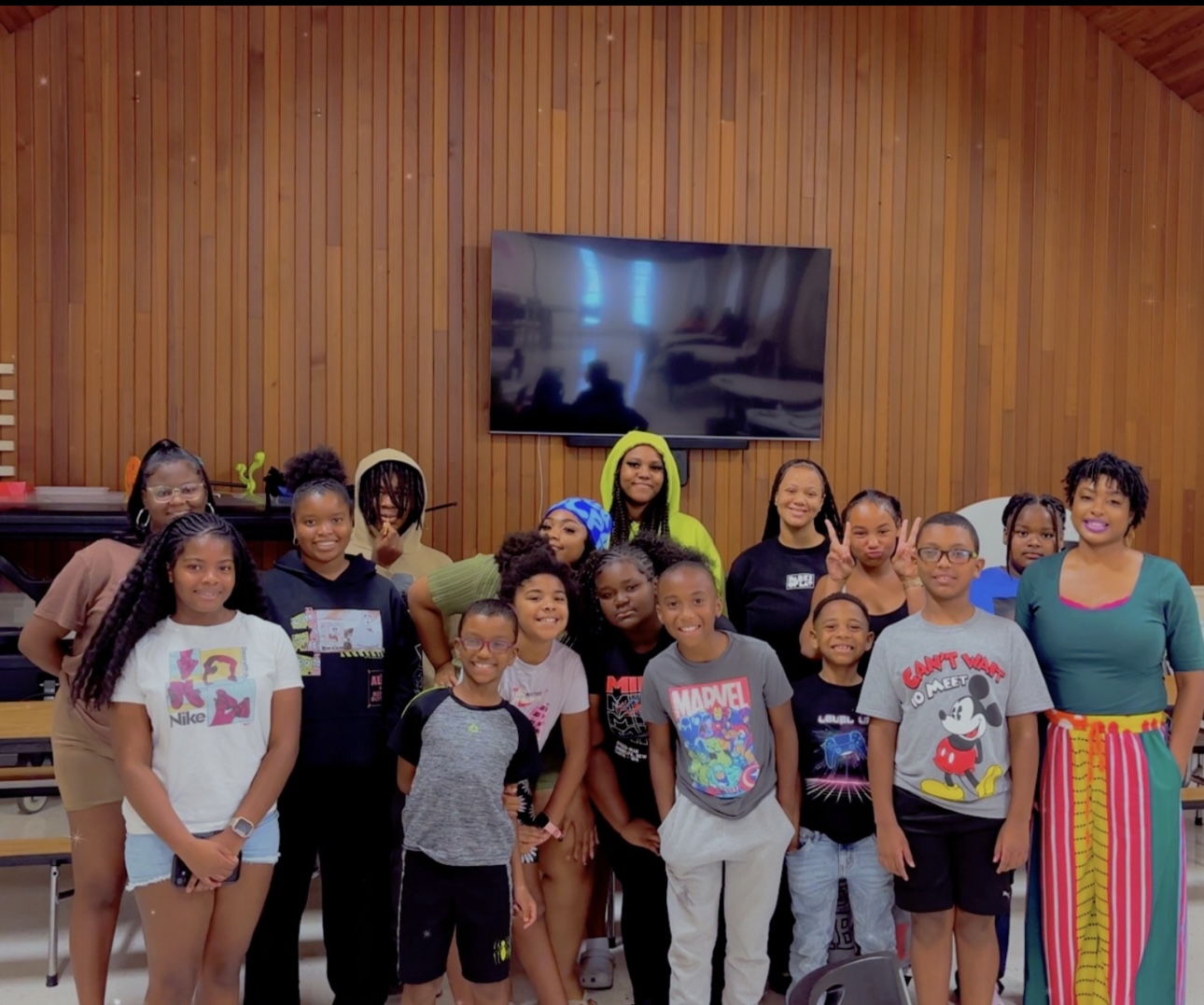 Group of children and two adults standing in front of a wooden wall with a TV screen, smiling and posing for a photo.
