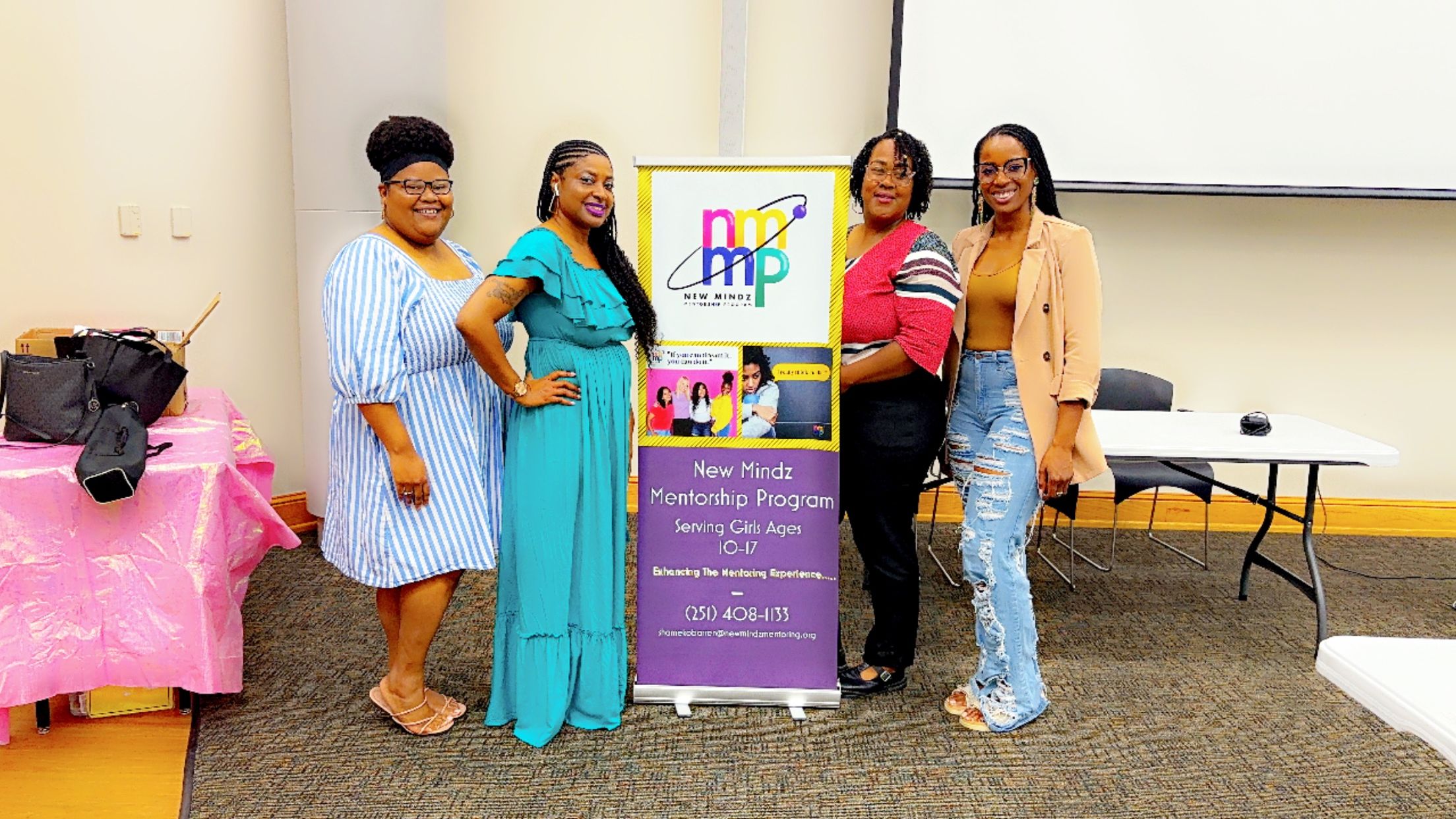 Three women standing in front of a purple banner, one holding a microphone, in a room with a table and a projector screen.