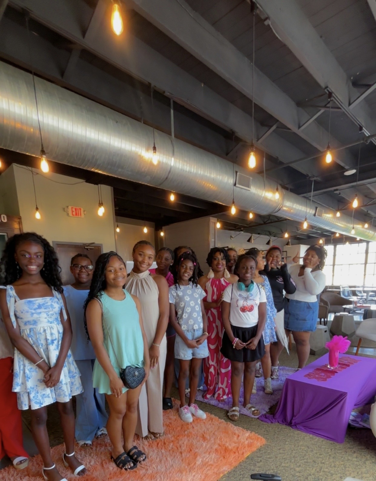 Group of children standing indoors near a purple table, with hanging lights and industrial ceiling visible, some smiling and posing.