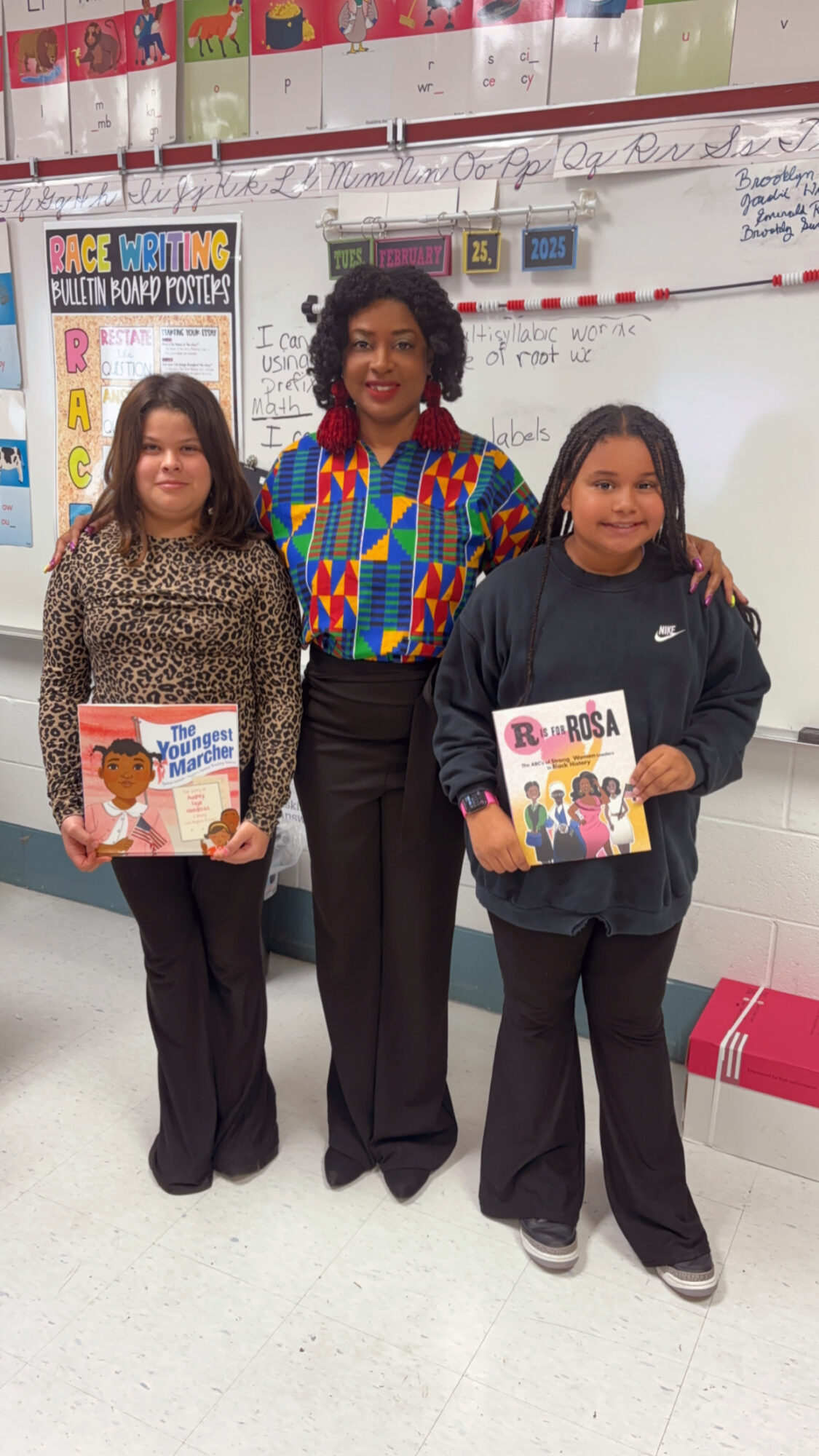 Three people standing in a classroom, two children holding books, and a woman in the middle smiling.