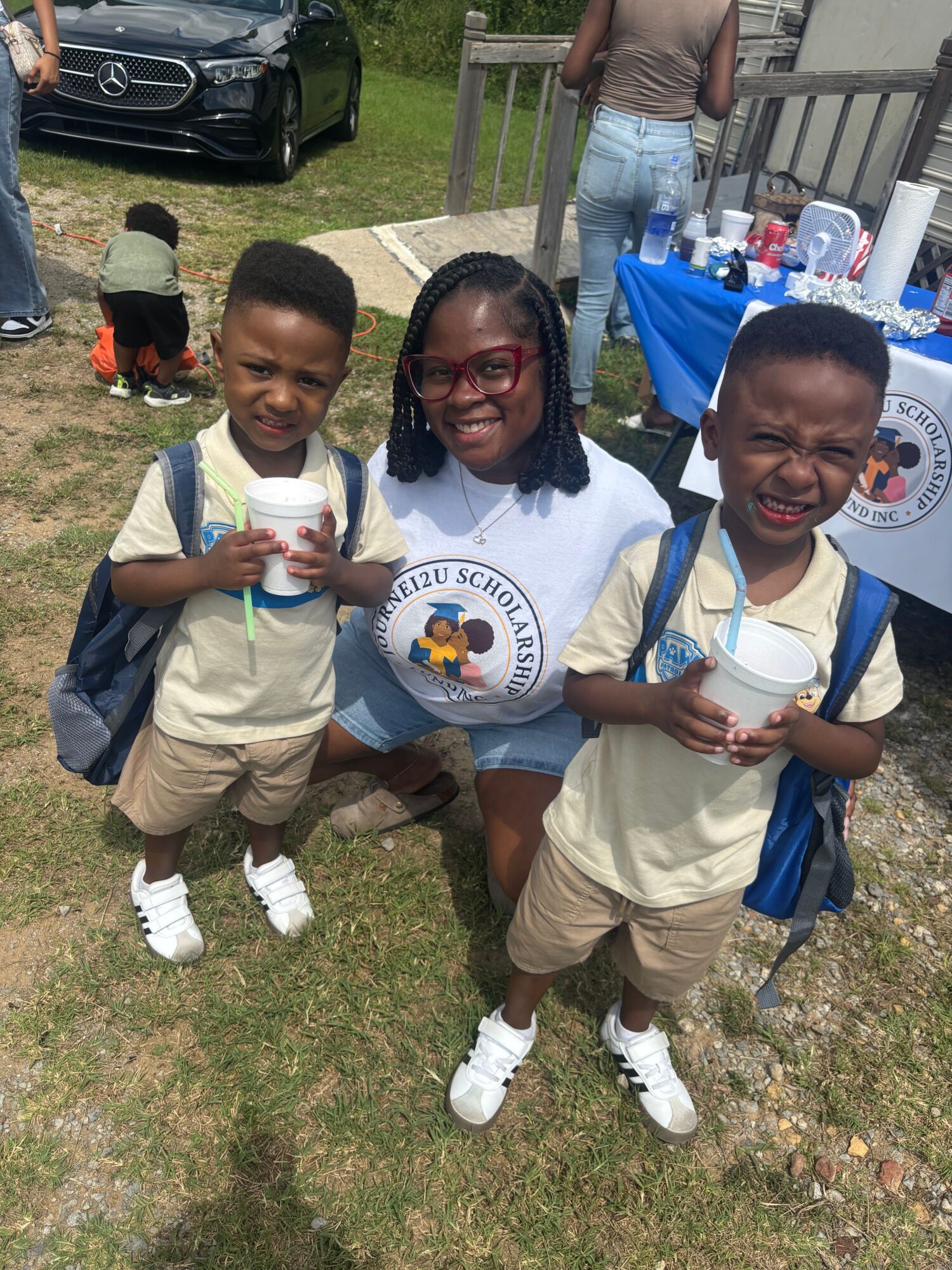 Three children in school uniforms holding drinks outdoors, smiling, with a person and a vehicle in the background.