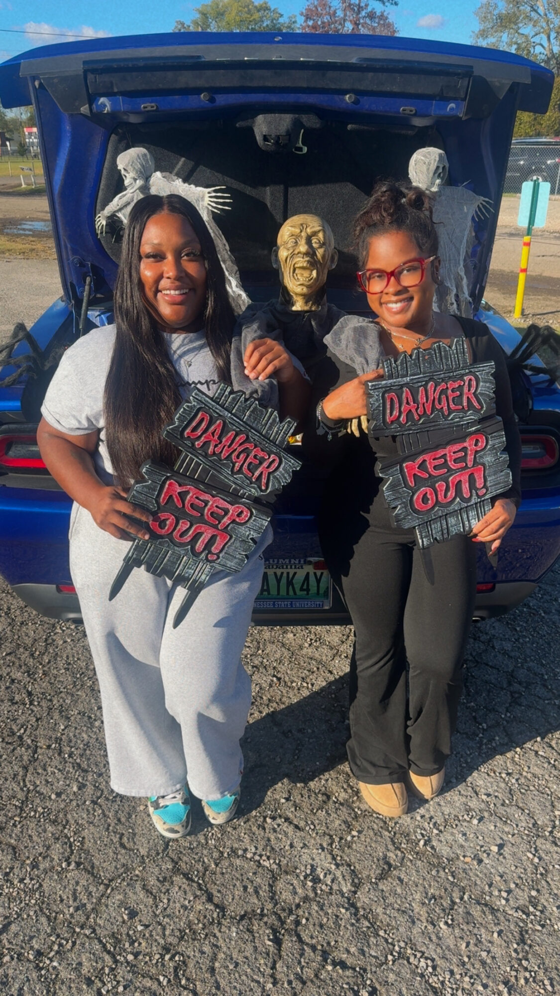 Two women holding signs that say 'DANGER KEEP OUT' stand in front of a car with its trunk open, a skull decoration inside, outdoors.