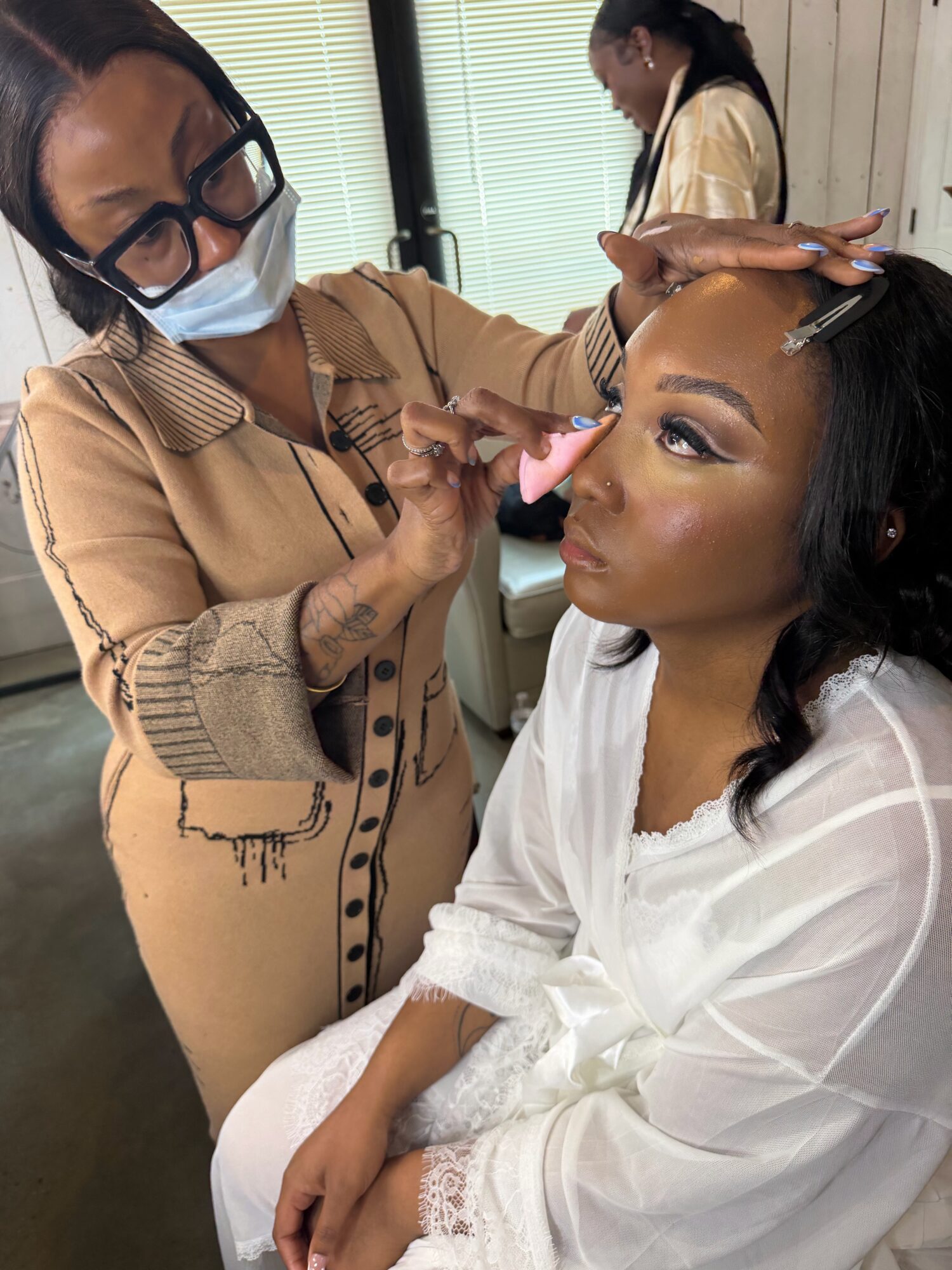 Makeup artist applying makeup to a woman seated, with another person in the background. Indoor setting with natural light.