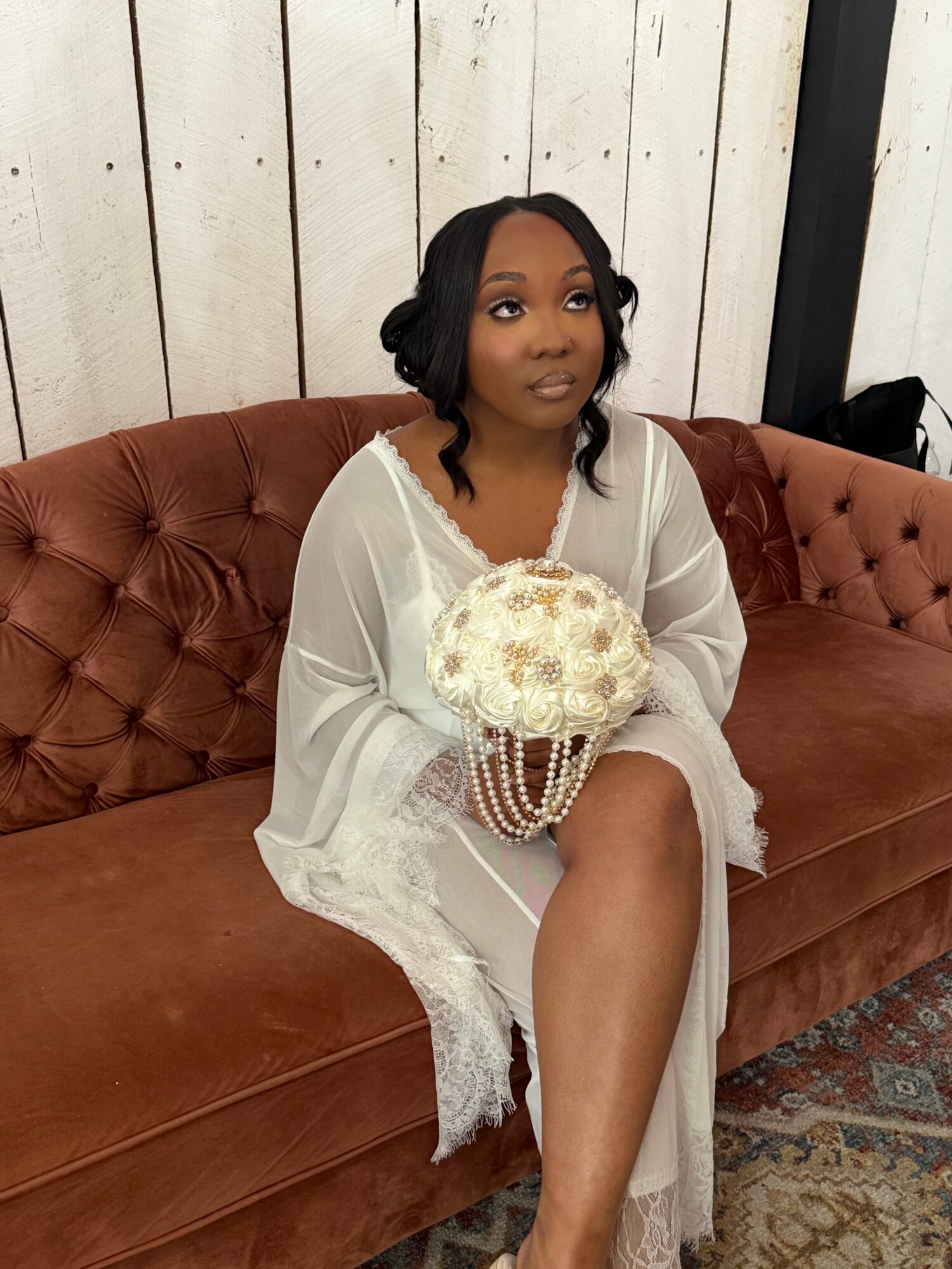 Woman in white dress holding a bouquet, sitting on a brown tufted sofa against a white wooden wall.