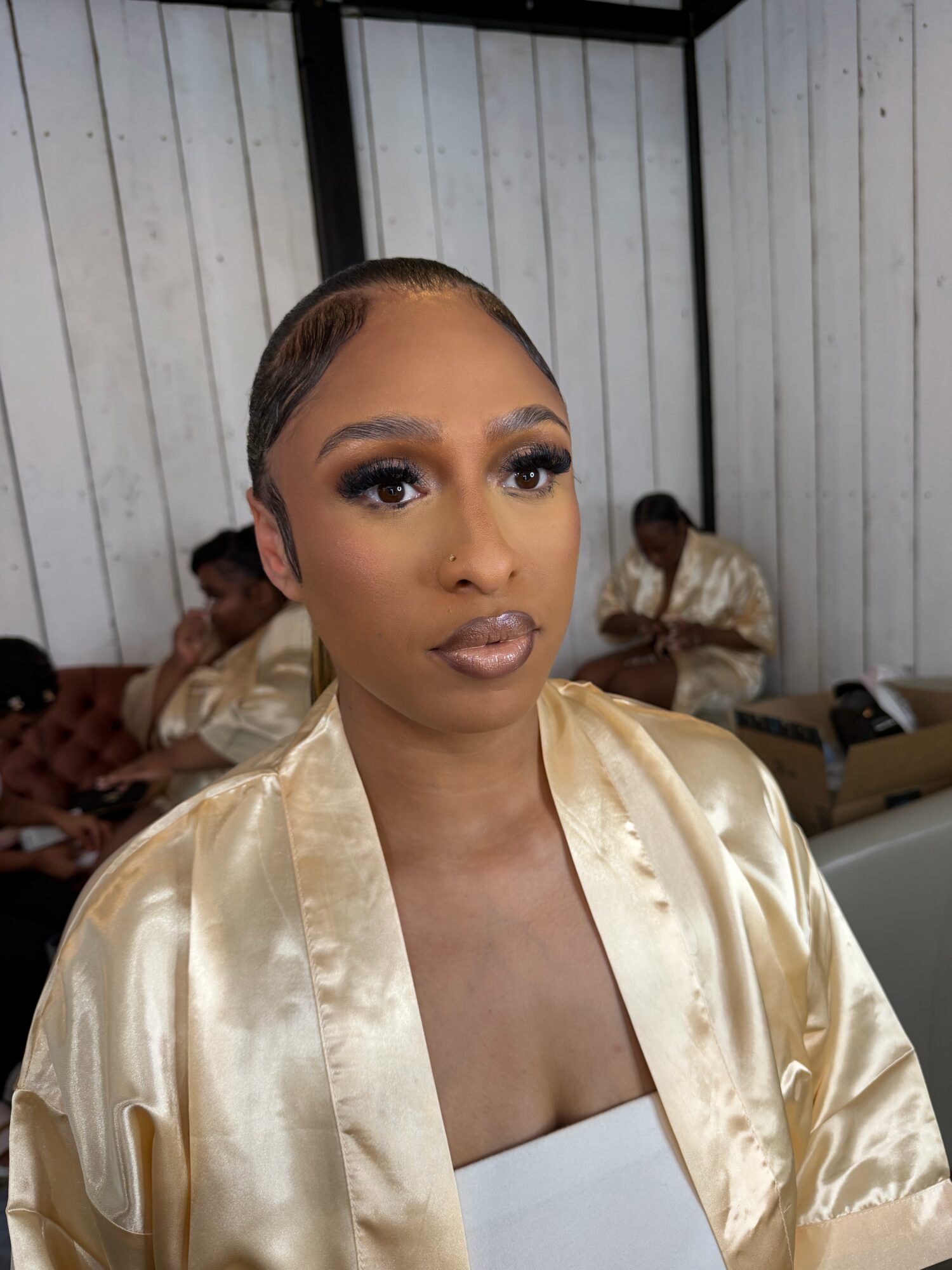 Woman with makeup and short hair in a cream satin robe, standing indoors with people in the background.
