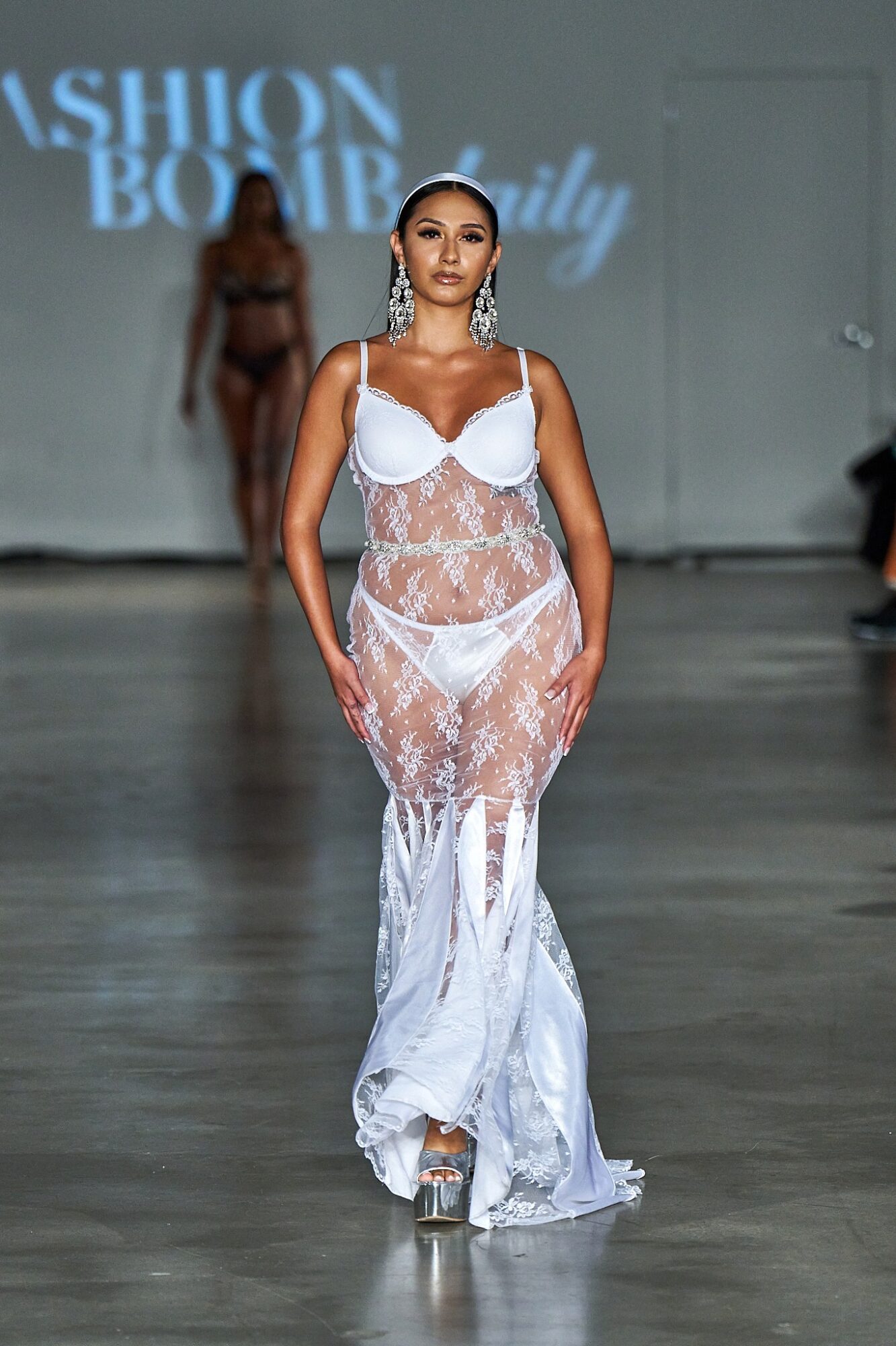 Model walking on runway in white lace dress with sheer panels, under a sign that reads 'Fashion Body'.