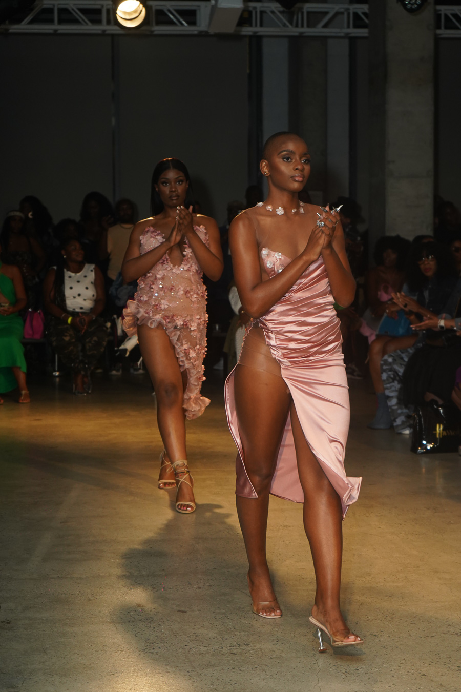 Two women walk on runway in elegant dresses, with audience in background, dim lighting, and spotlight on models.