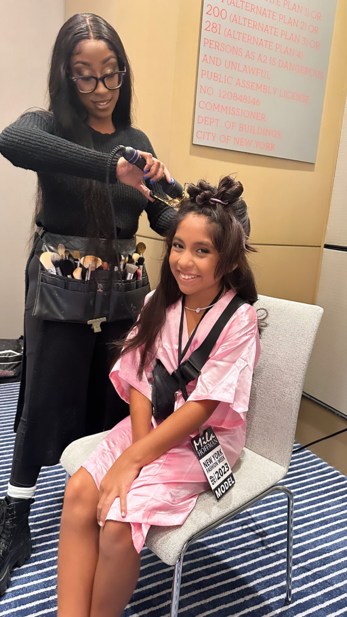 A woman styles a young girl’s hair in a salon, girl sits on a chair wearing a pink gown, smiling.