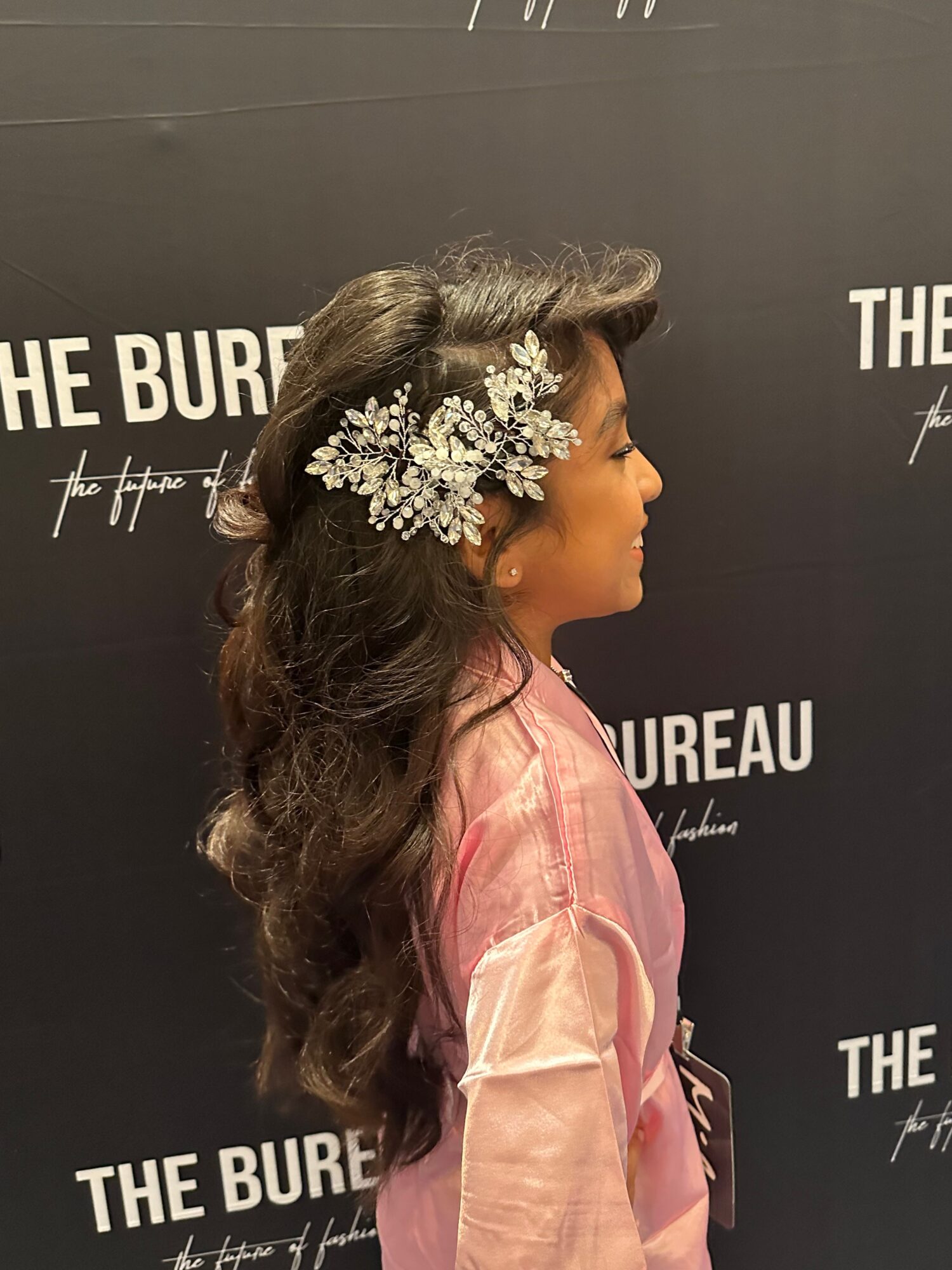 Side view of a woman with long dark hair and floral hair accessory, wearing a pink satin dress, against a black background.