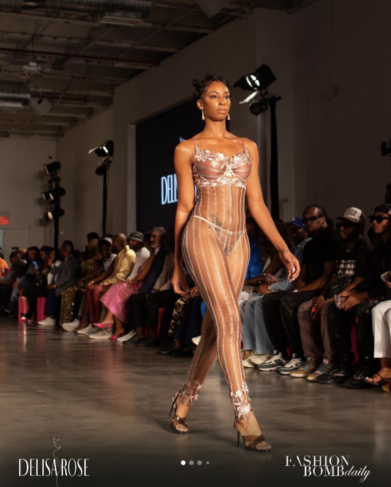 Model walking on runway in sheer, striped outfit with floral top, audience seated on both sides, fashion show setting.