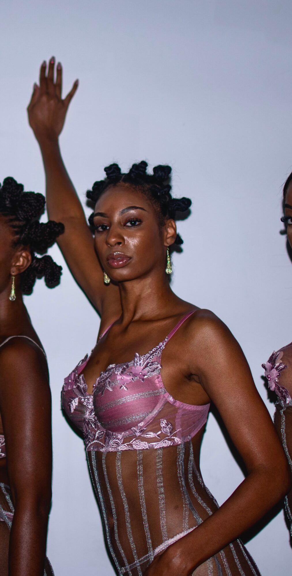 Woman with styled hair and makeup, wearing a pink and brown corset, raising her right arm, against a plain background.