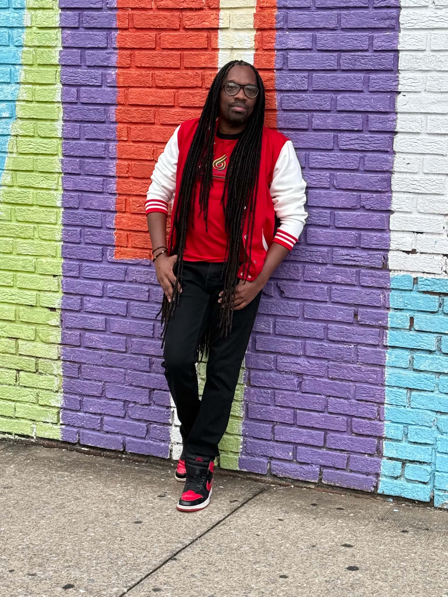 Person with long hair and glasses standing in front of a colorful brick wall, wearing a red shirt, white and red jacket, black pants, and red shoes.