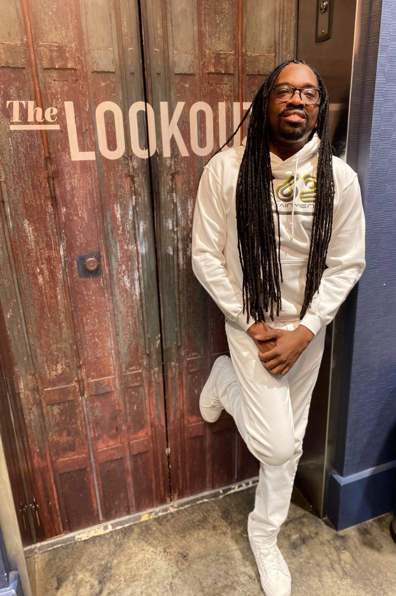 Man with long dreadlocks and glasses stands with hands clasped in front of wooden door with 'The LOOKOUT' sign.