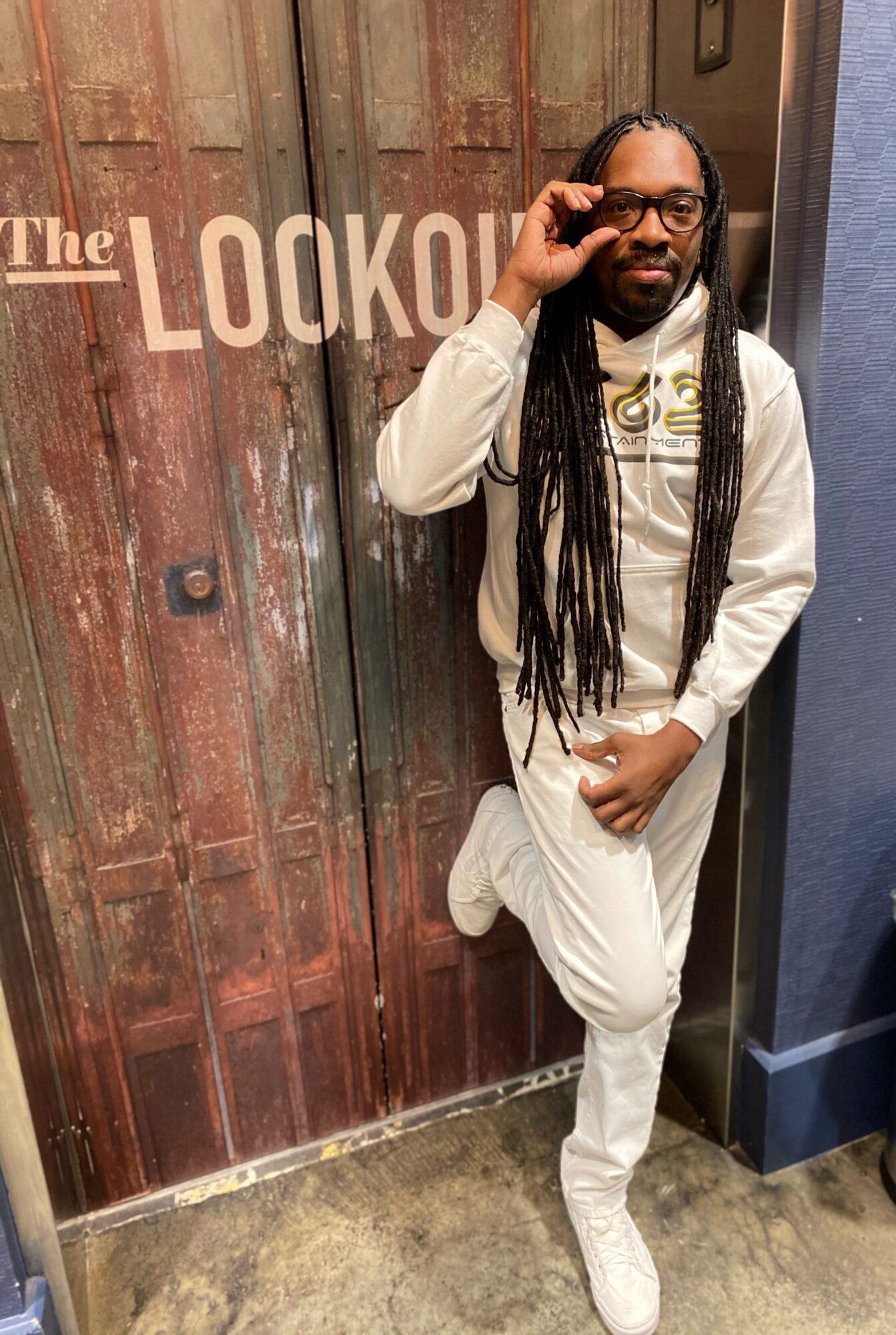 Person with long dreadlocks and glasses leaning against a wooden door with 'The LOOKOUT' sign, wearing white outfit, adjusting glasses.