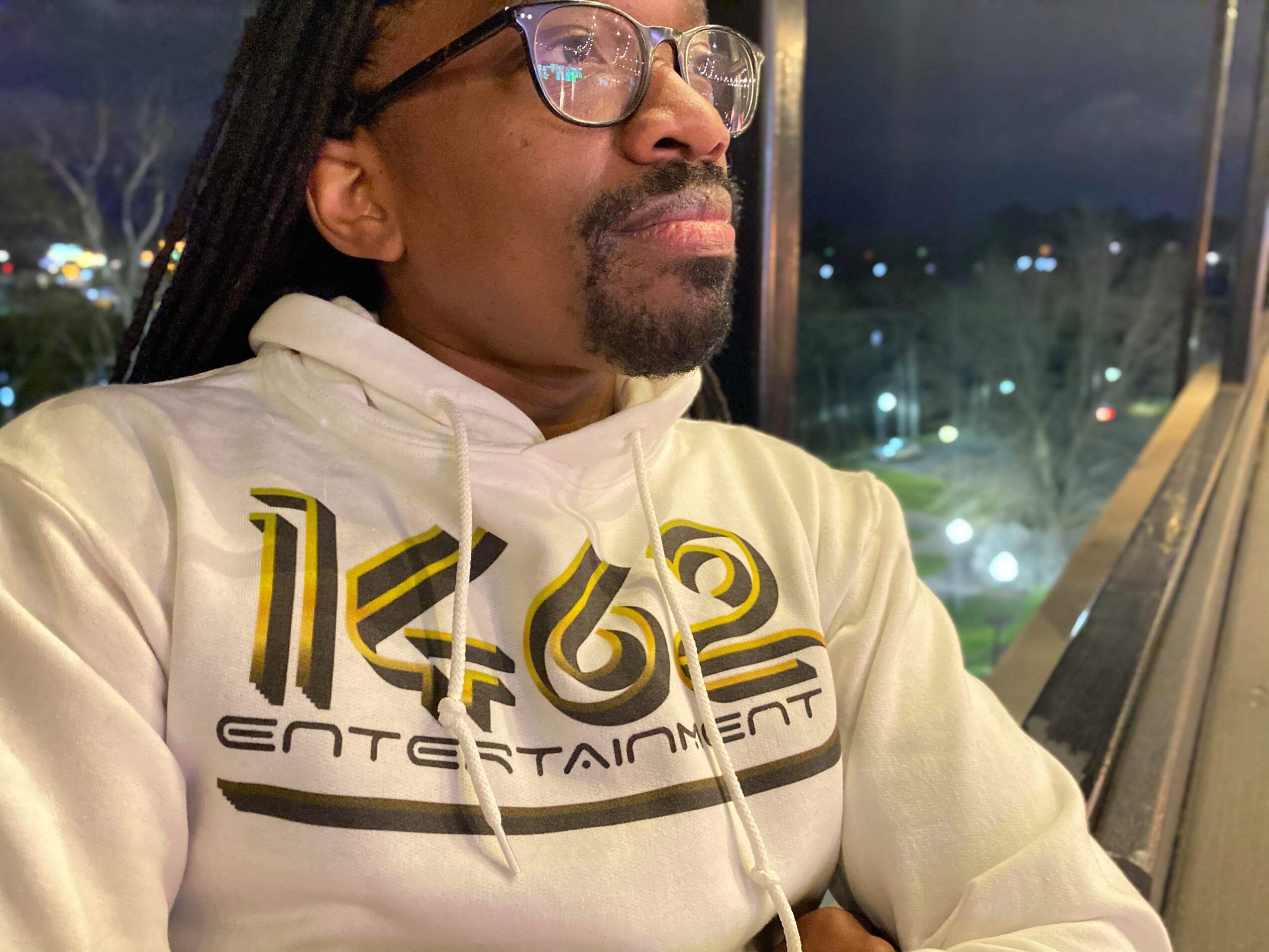 Man with glasses and long hair looking out a window at night, city lights visible outside.