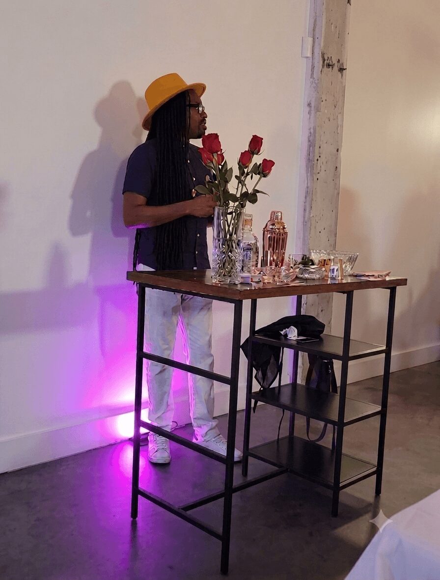 Person with long hair, yellow hat, and glasses holding roses, standing near a table with drinks and glasses, against a white wall.