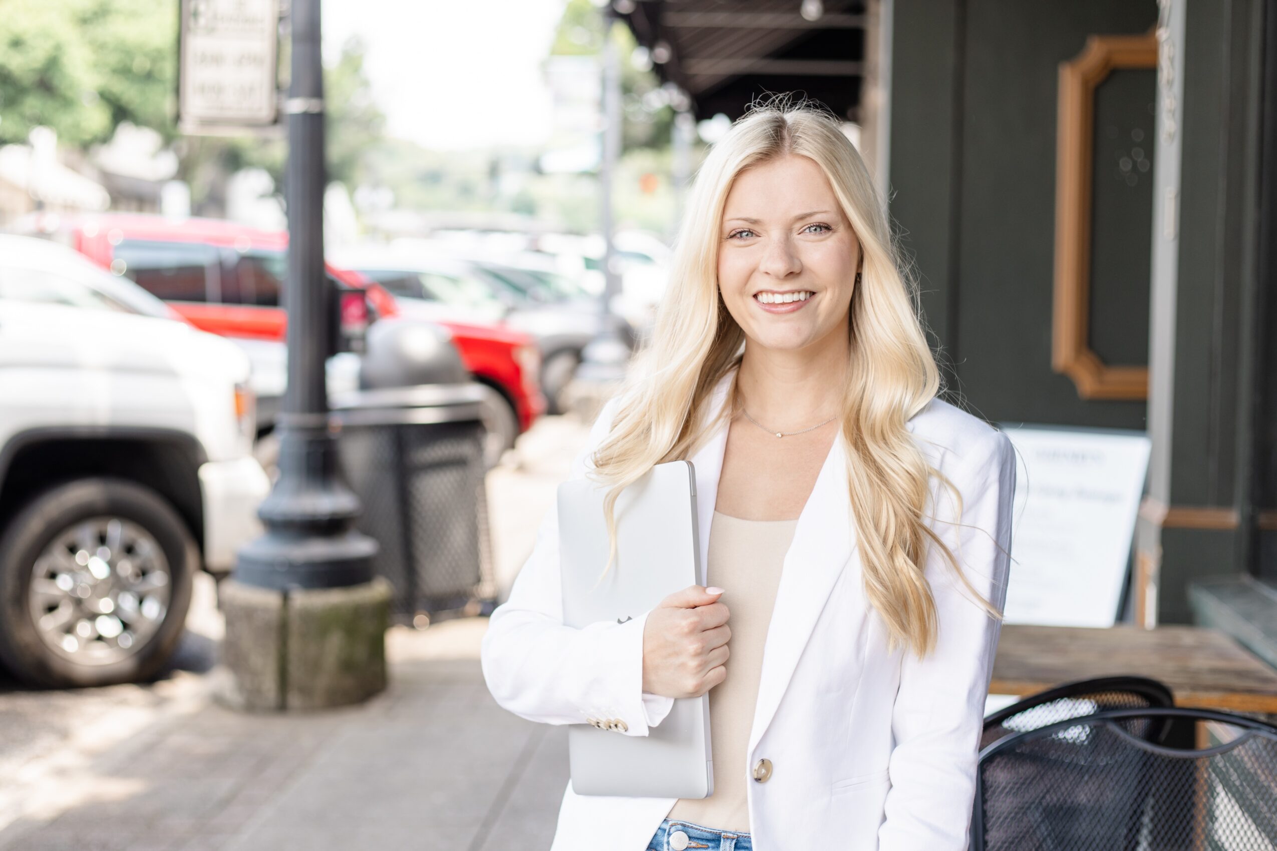 Smiling woman with long blonde hair standing outdoors on a city sidewalk, holding a white bag, wearing a white blazer.