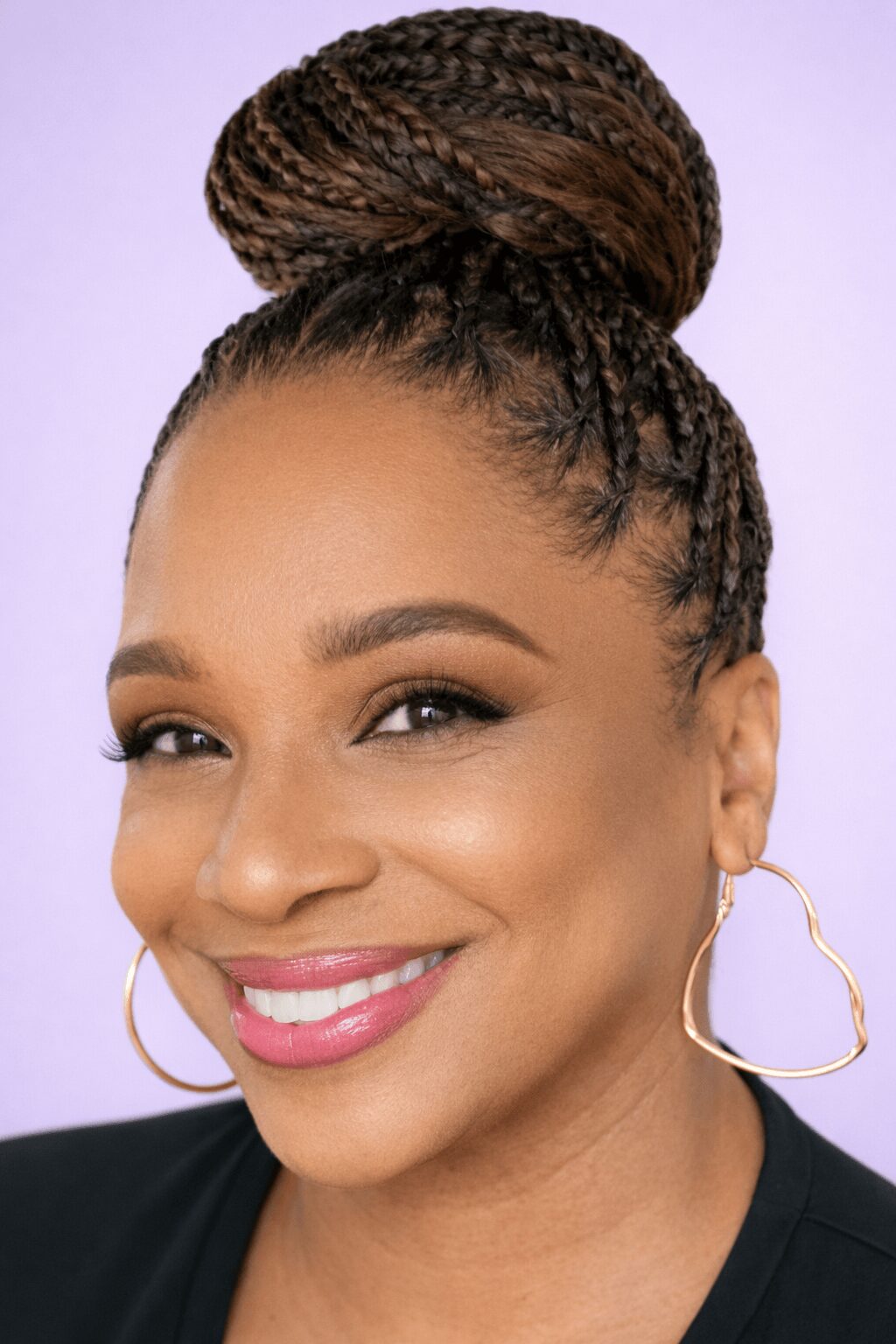 Close-up of a smiling woman with braided hair styled in a high bun, wearing hoop earrings and a black top.