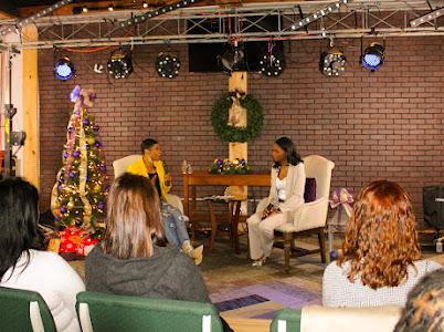 Two women sit on stage with a Christmas tree and decorations, audience seated in front, brick wall background.