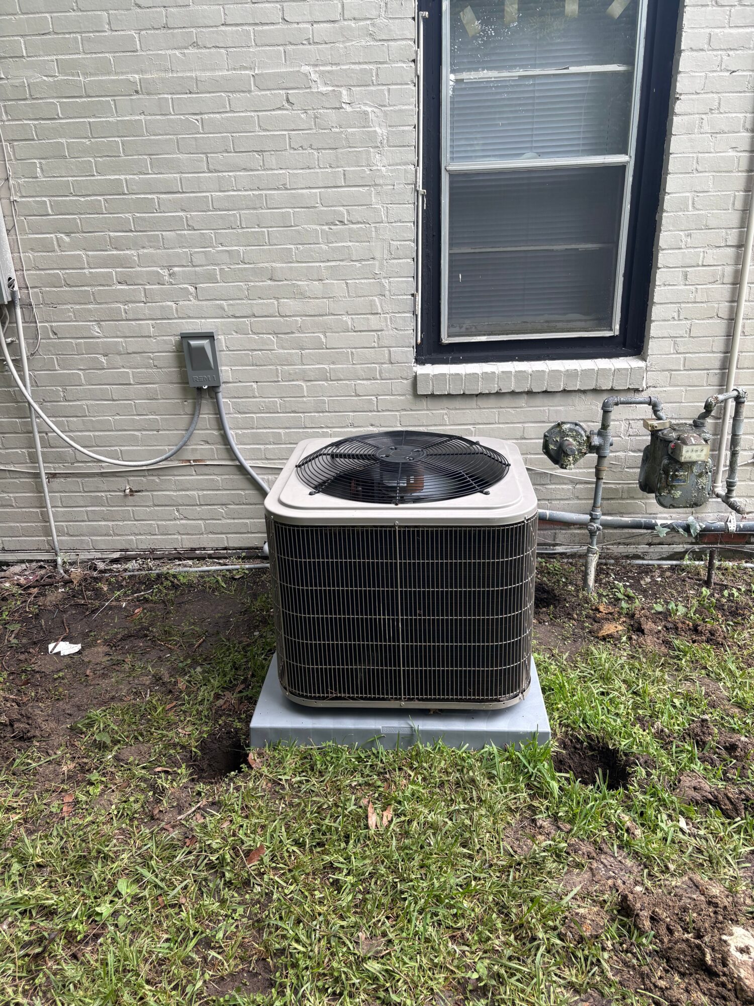 Outdoor air conditioning unit installed on a concrete pad next to a brick wall with a window, on grassy ground.