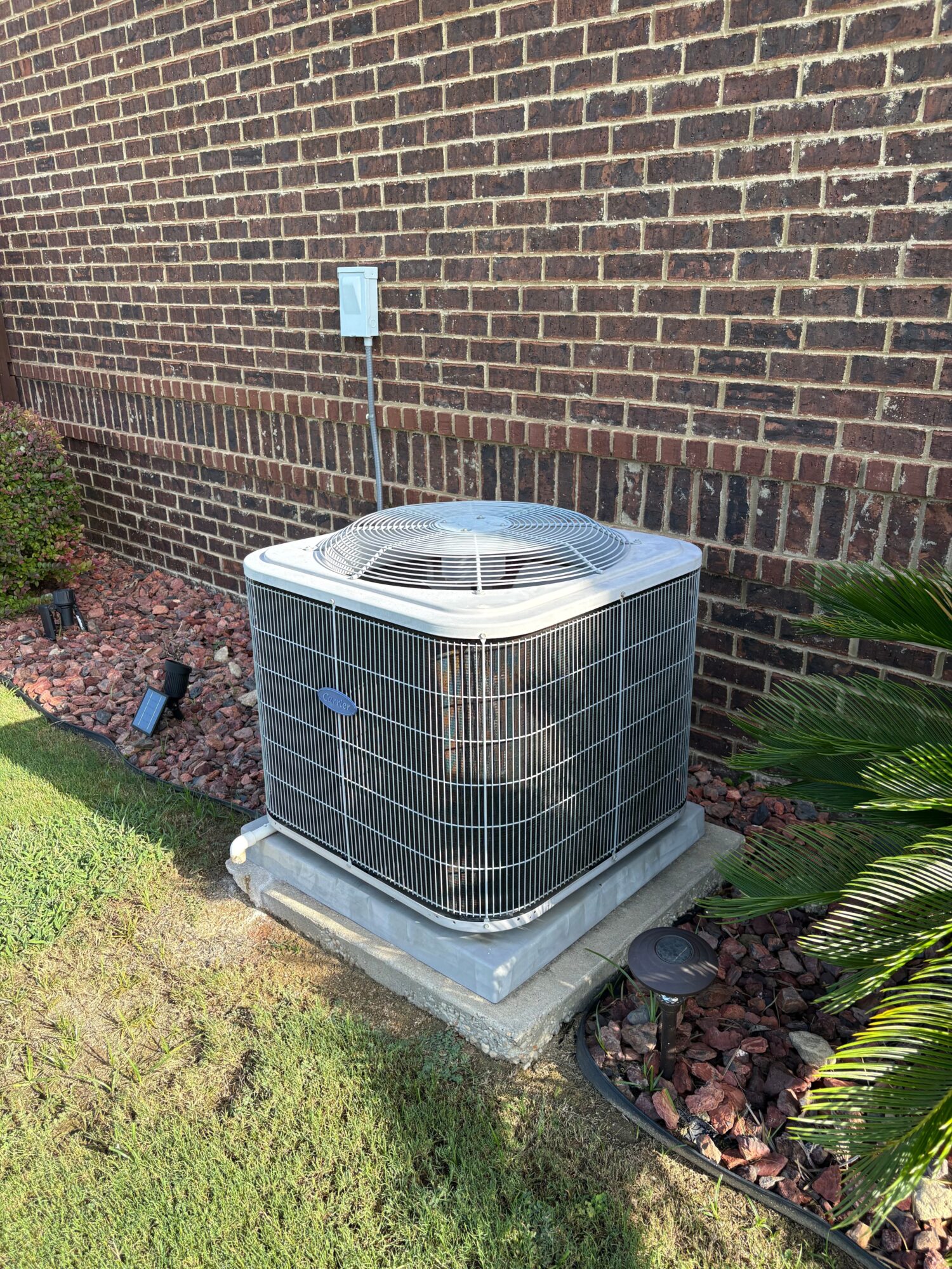Air conditioning unit installed on a brick wall with plants nearby, viewed from the side.