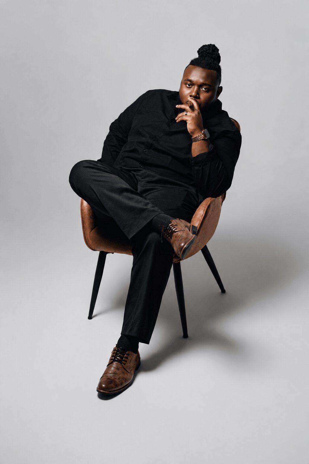 Person sitting on a chair with legs crossed, wearing black clothing and brown shoes, against a plain gray background.