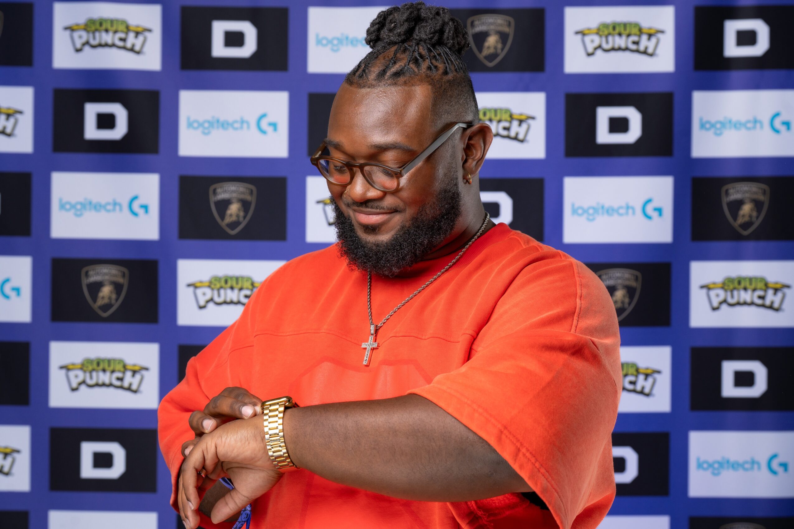 Man with glasses, beard, and braided hair in an orange shirt, standing in front of a blue and black patterned backdrop.