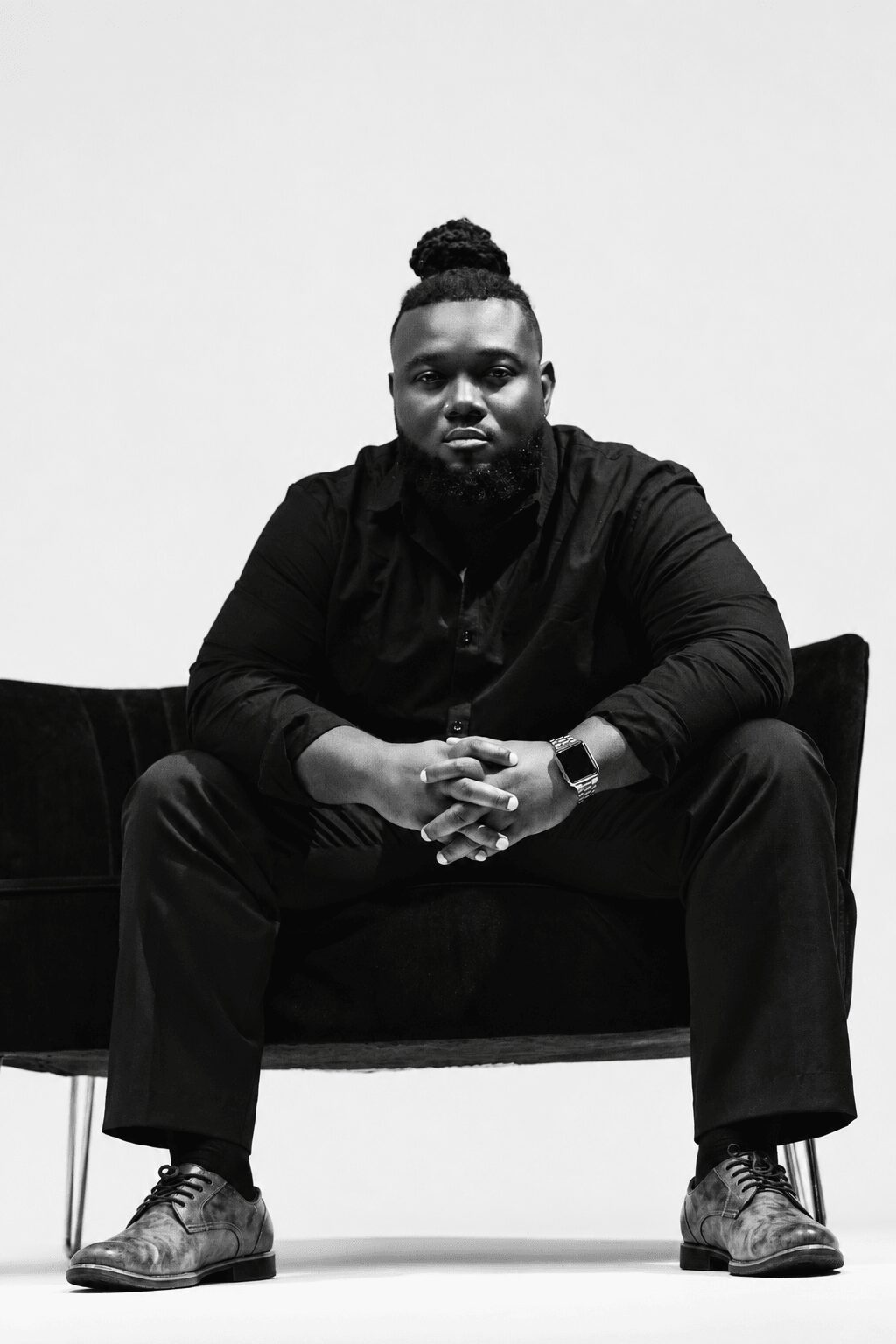 Man with a bun hairstyle sitting on a dark sofa, hands clasped, wearing dark clothing and shoes, against a plain background.