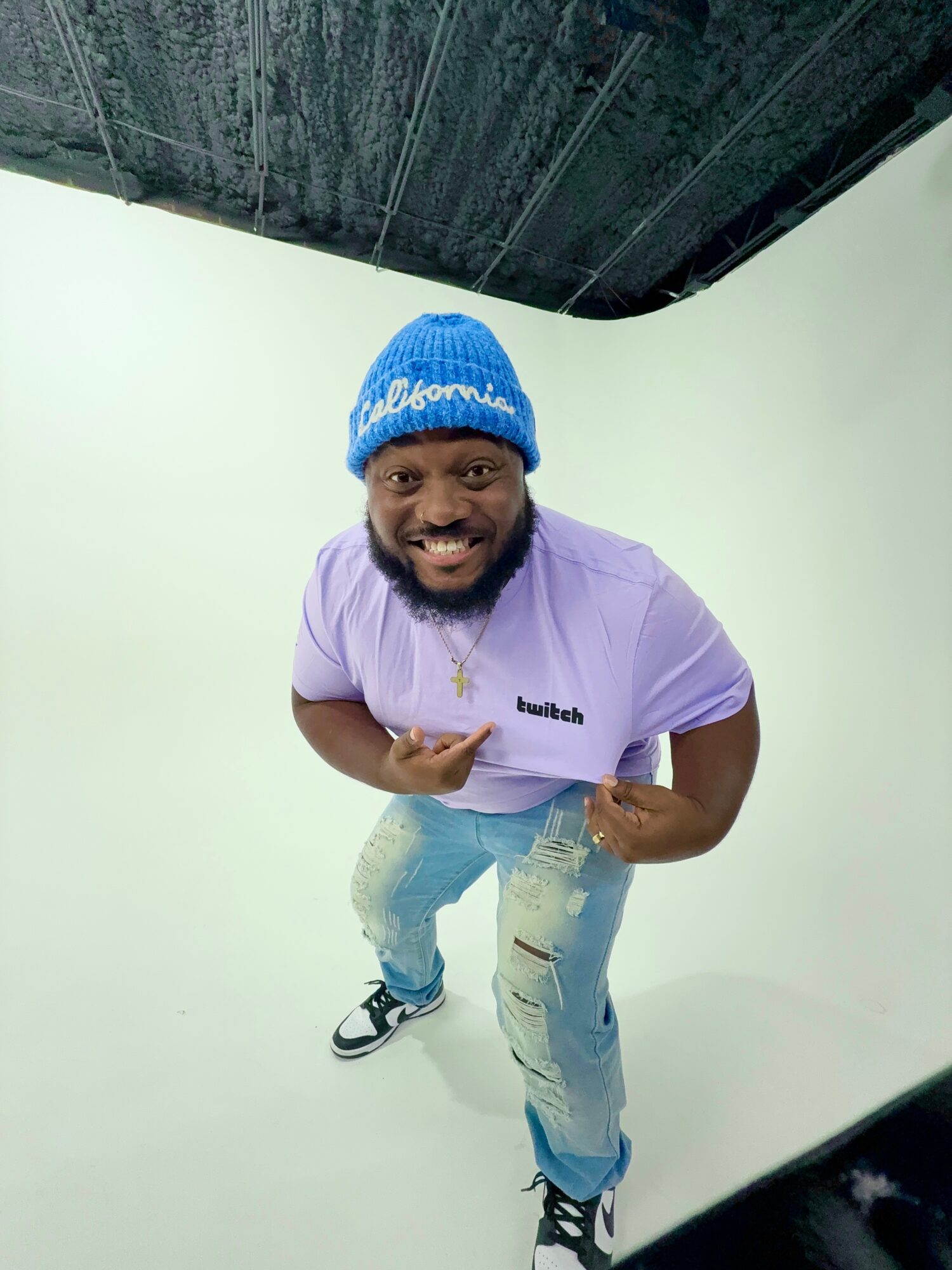 Man smiling, wearing a blue beanie, purple shirt, and jeans, pointing with both hands, standing in a white studio.