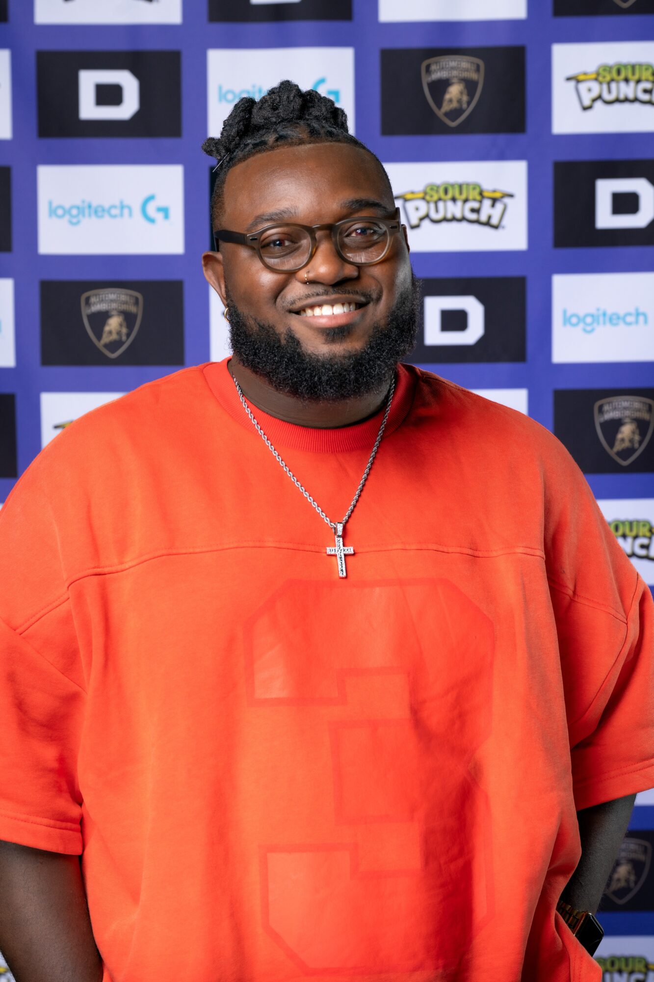 Man with glasses, beard, and a cross necklace smiling in front of a blue and black backdrop with logos.