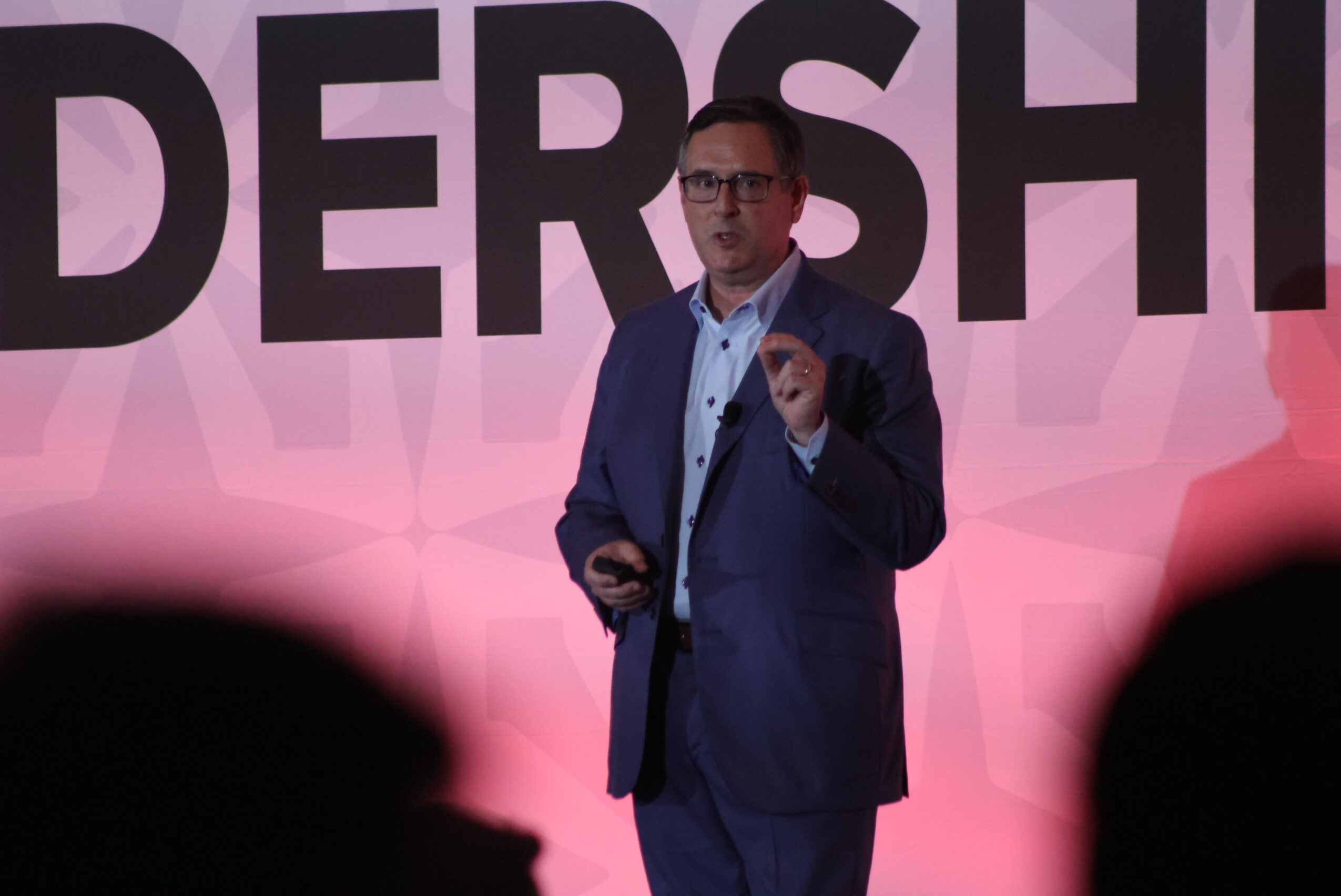 Man in a suit speaking on stage with large text behind him, pink background, holding a device, audience in foreground