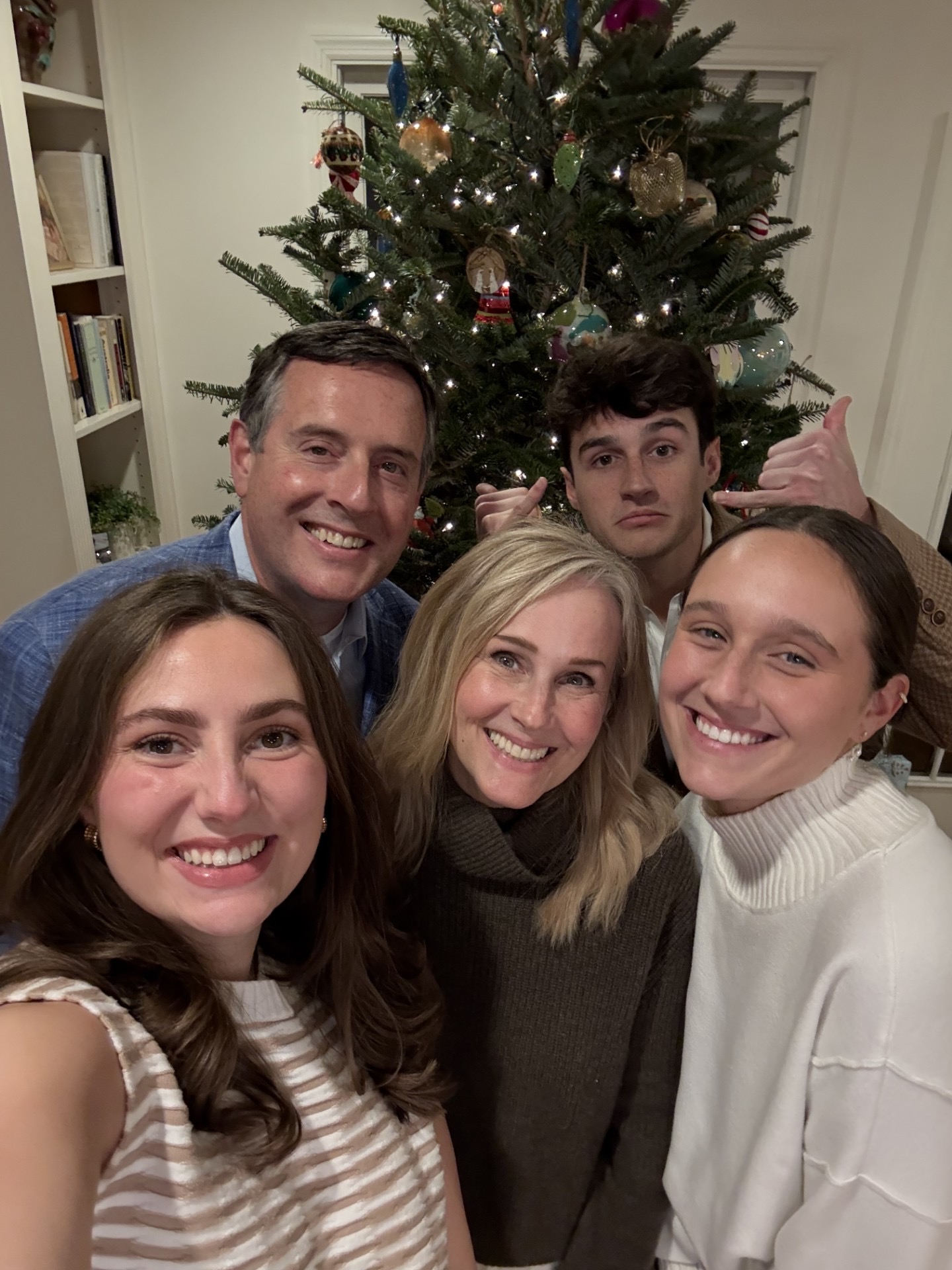 Five people smiling in front of a decorated Christmas tree, indoors, with bookshelves in the background.