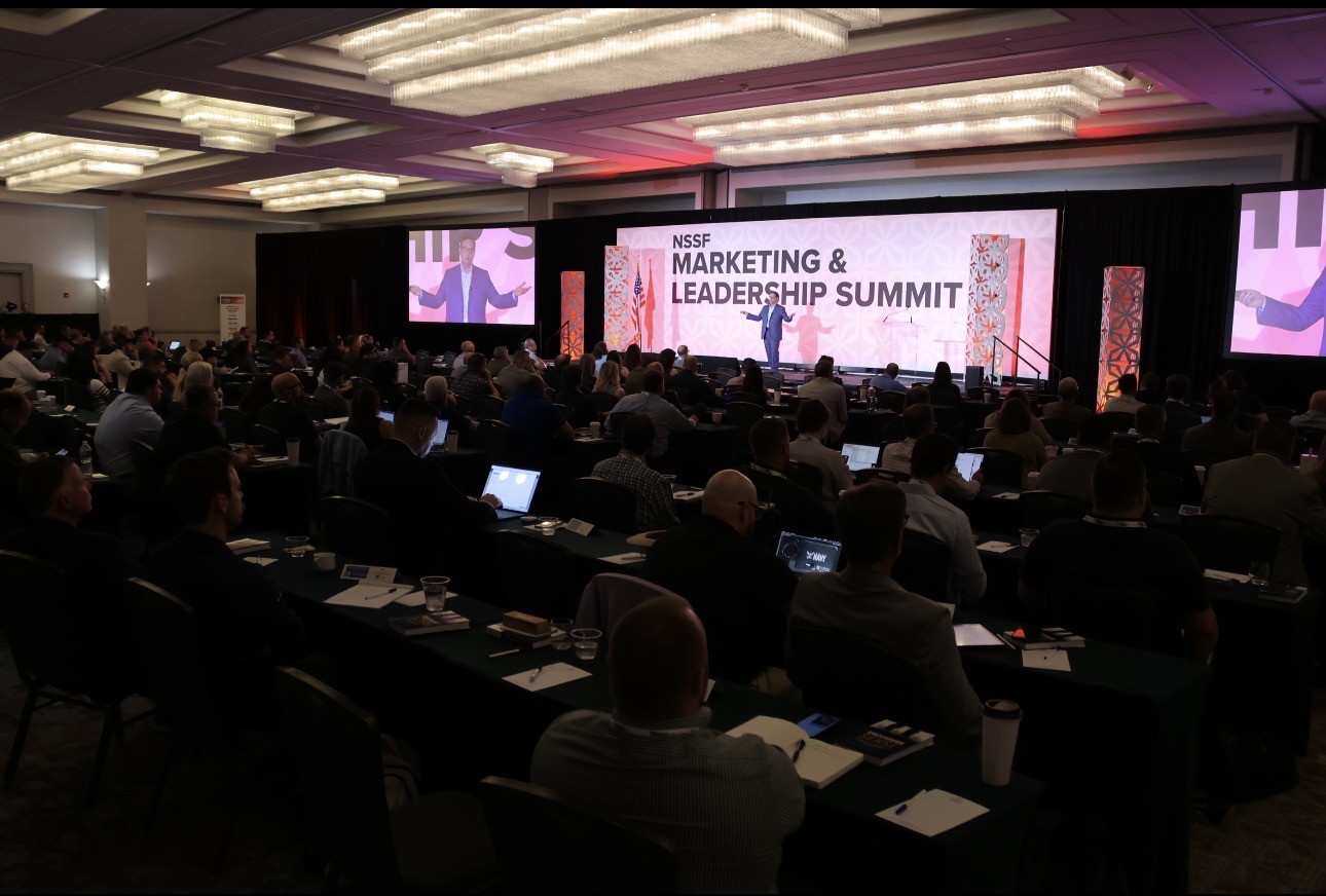 Conference room with audience watching a speaker on stage at the Marketing & Leadership Summit, large screens display event name.