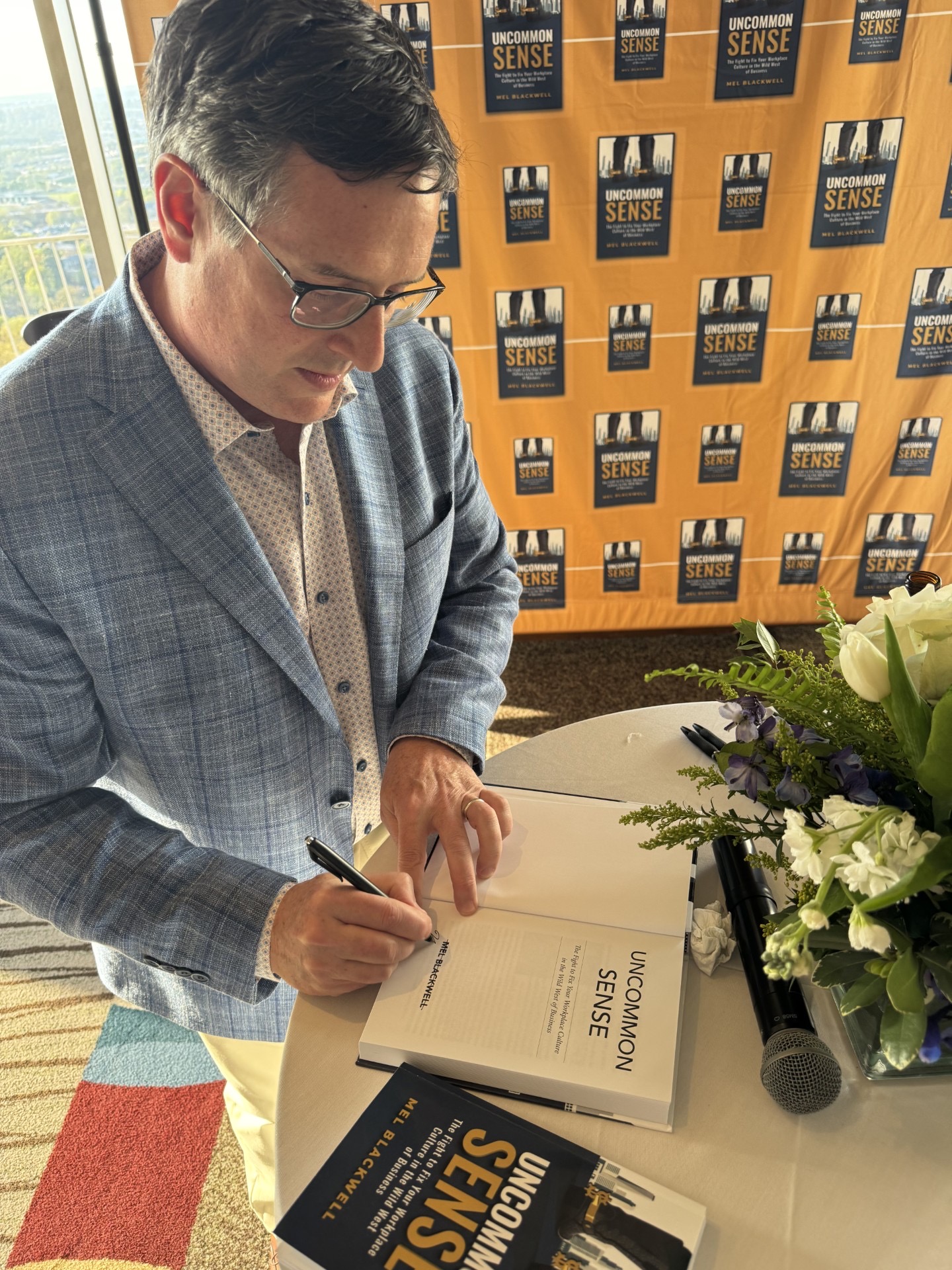 Man in glasses and checkered blazer signing a book at a table with a bouquet of flowers and a microphone.