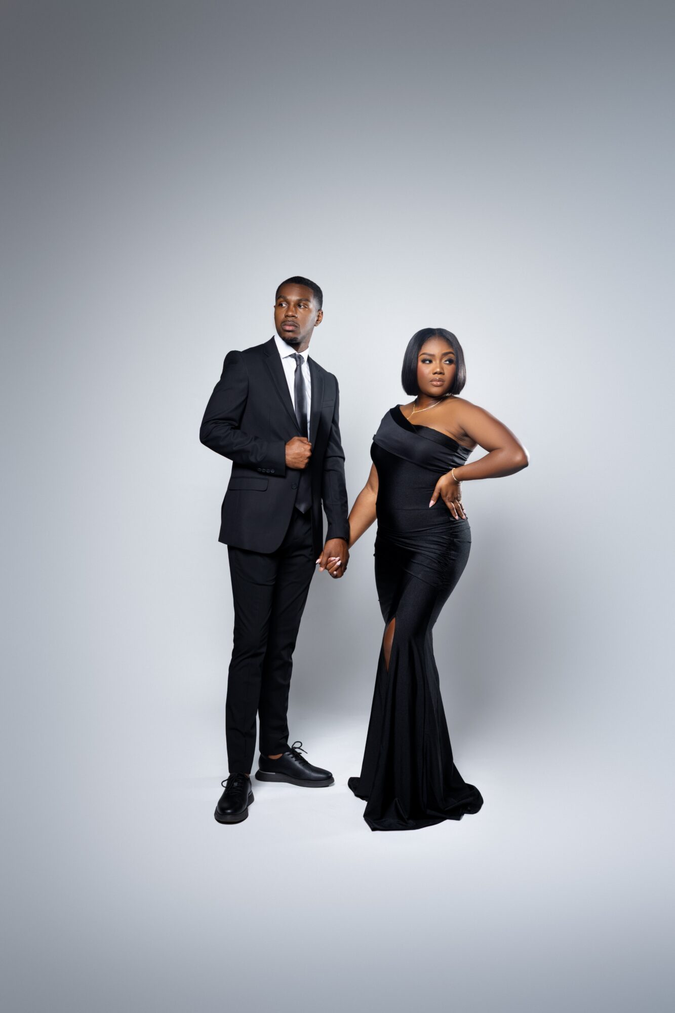 A man and woman in formal attire stand against a plain background, with the woman in a black gown and the man in a suit.