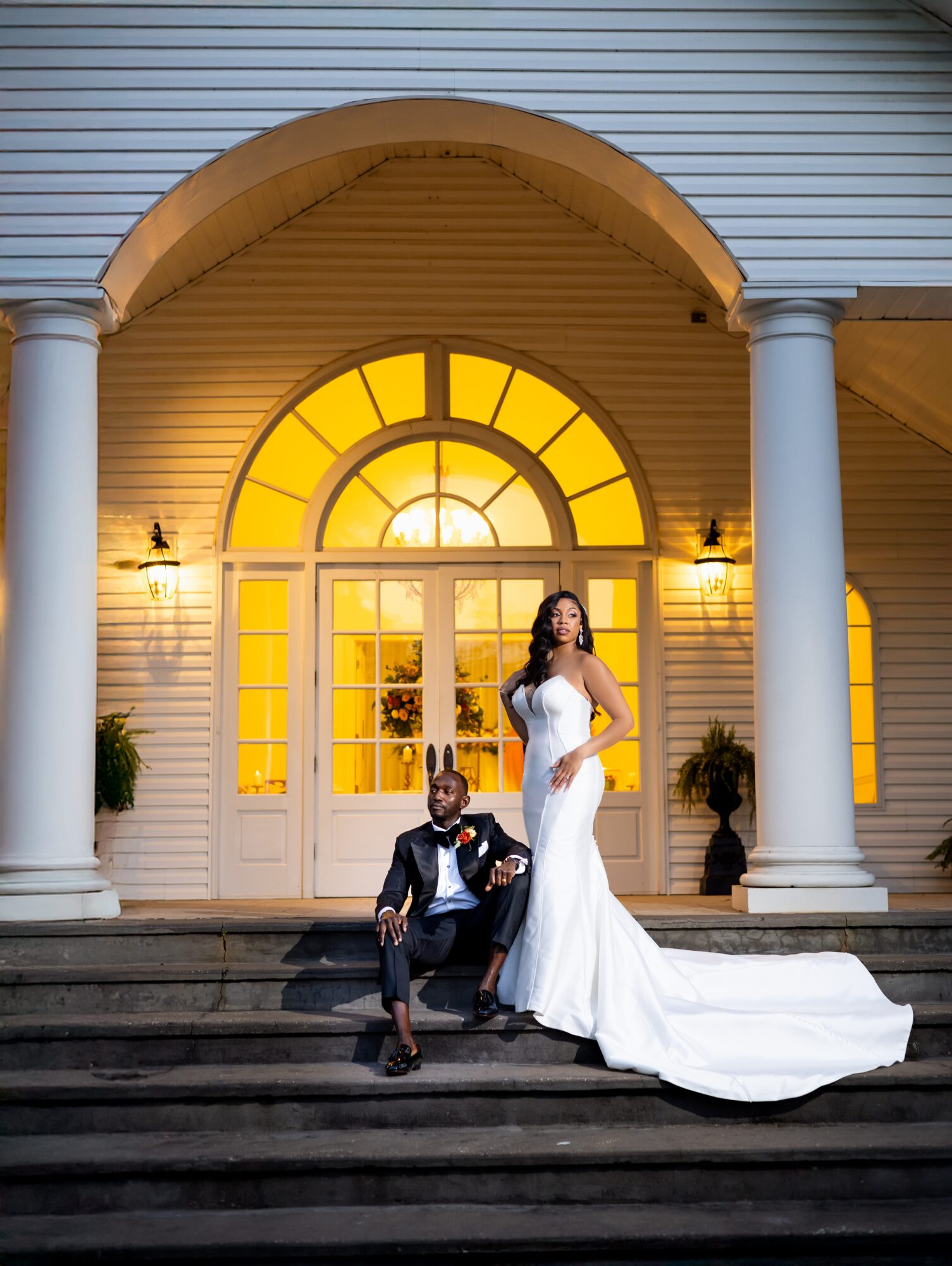 A bride in a white wedding dress and a groom in a suit pose on steps in front of a house with large arched window and columns.