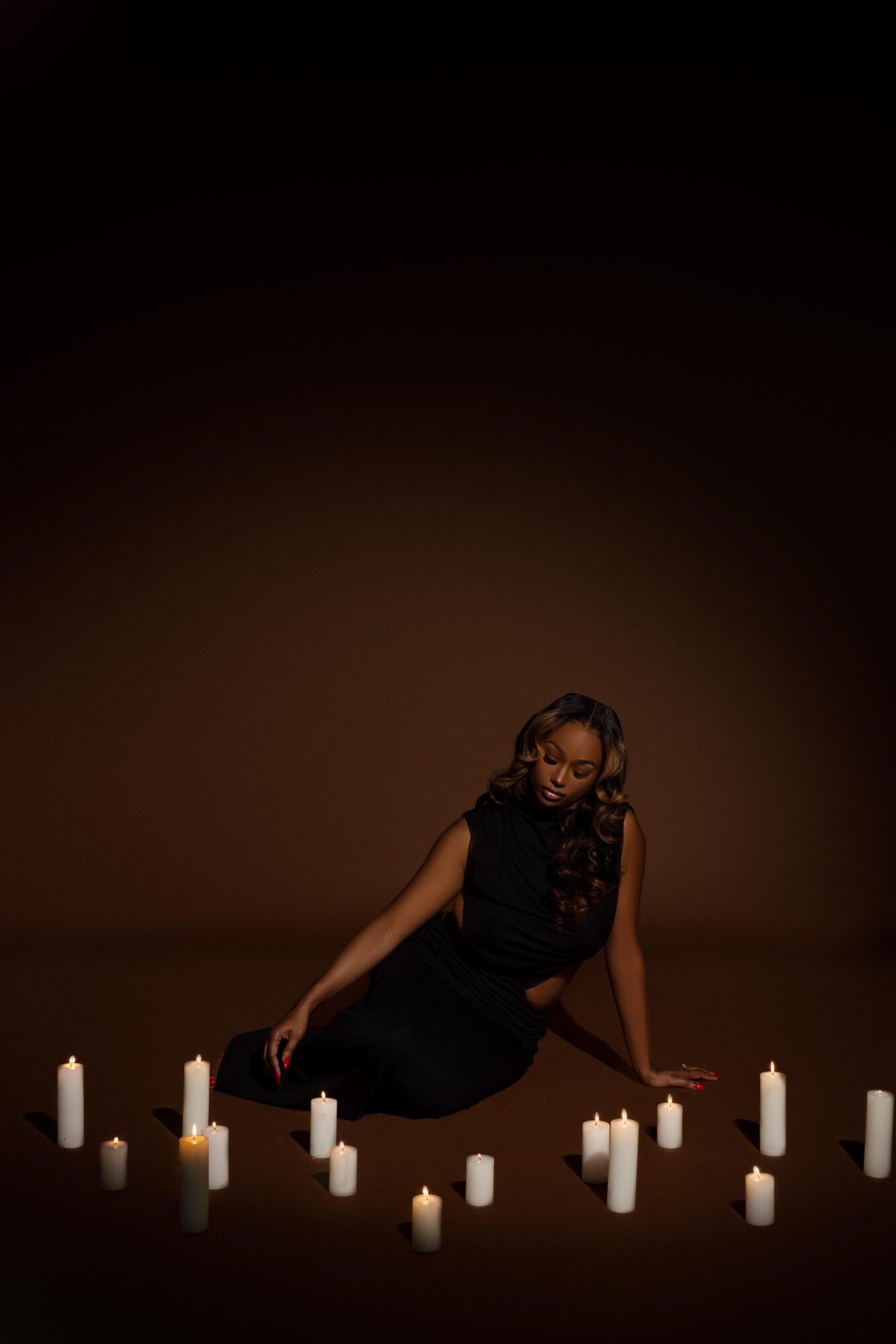 Woman in black dress sitting among lit candles on dark background, head tilted downward, smiling.