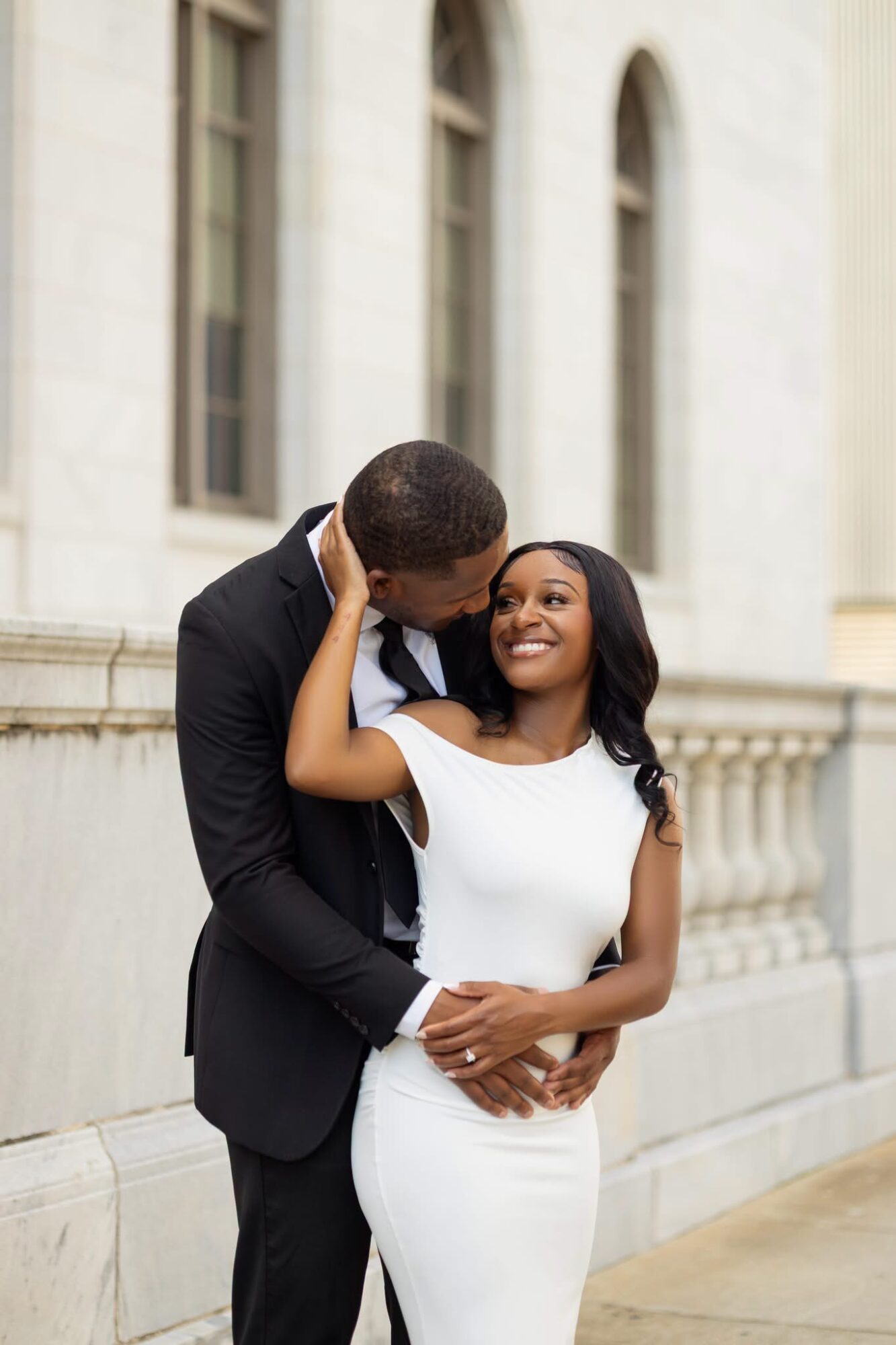 A couple stands close together outside a building with large windows, smiling and embracing each other.