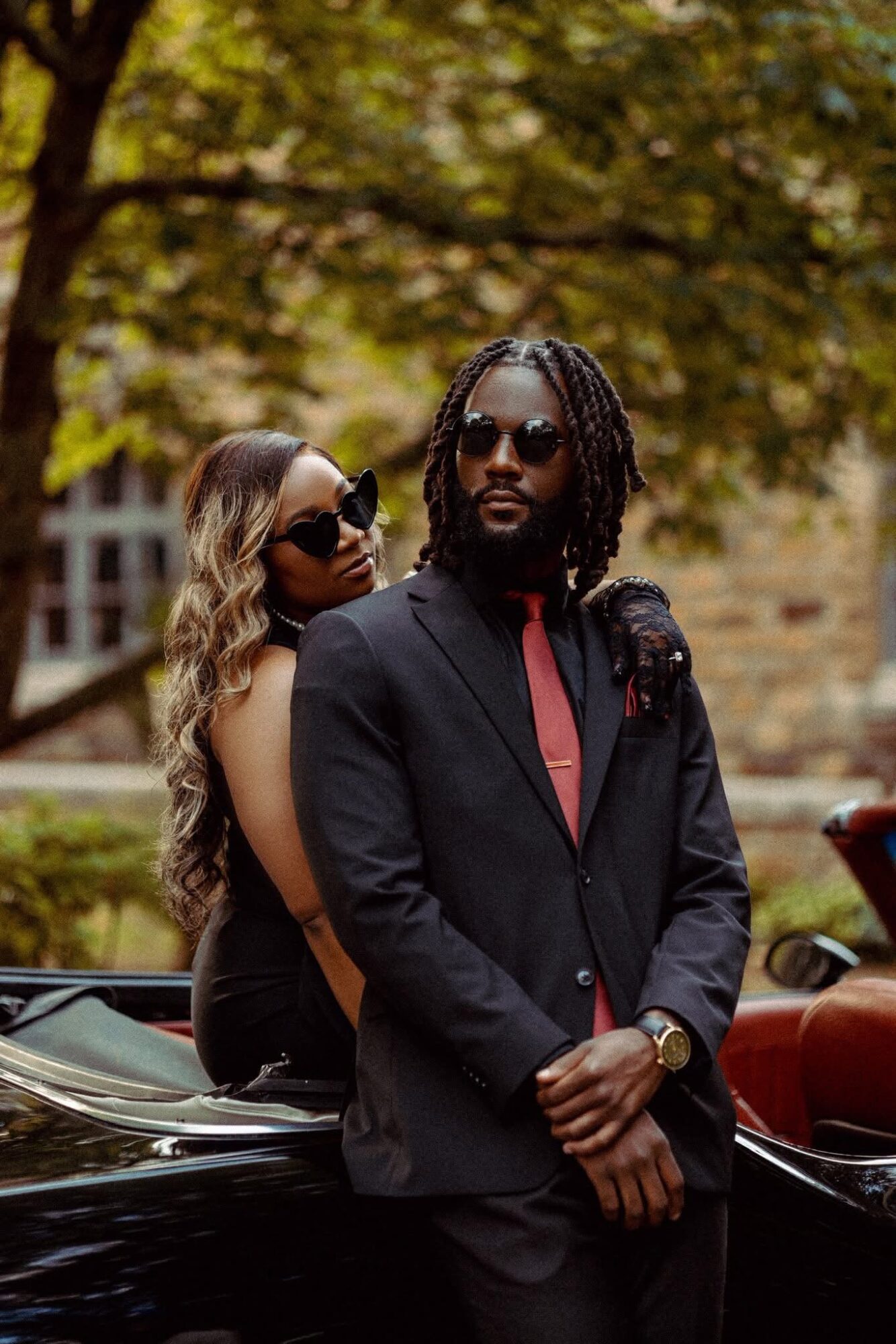 Two people, a man and a woman, stand outdoors with trees and cars in the background. The man wears sunglasses and a suit, the woman has long hair and sunglasses.