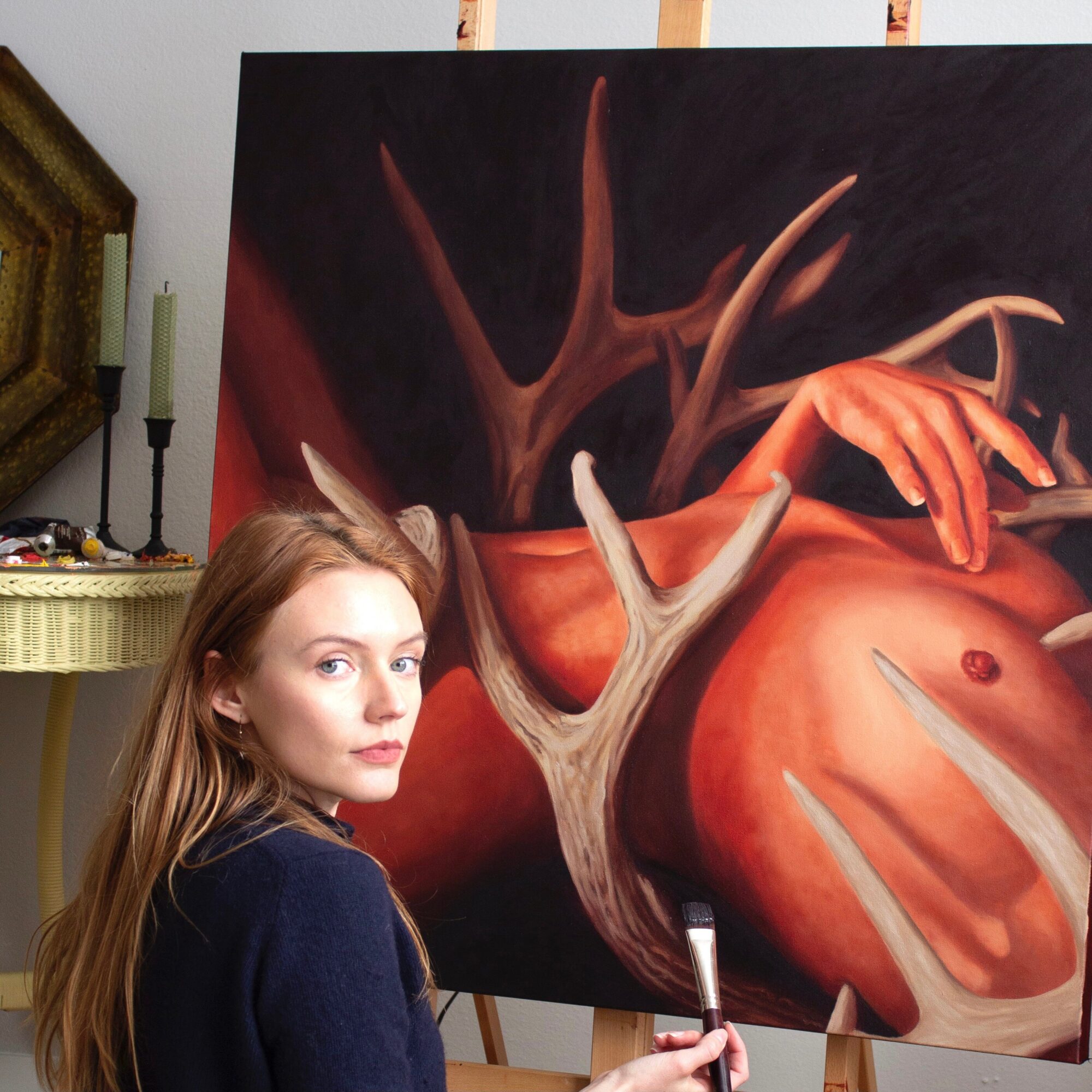 Young woman with long hair looking at the camera, standing next to a large painting of a surreal scene with a hand, antlers, and a body.