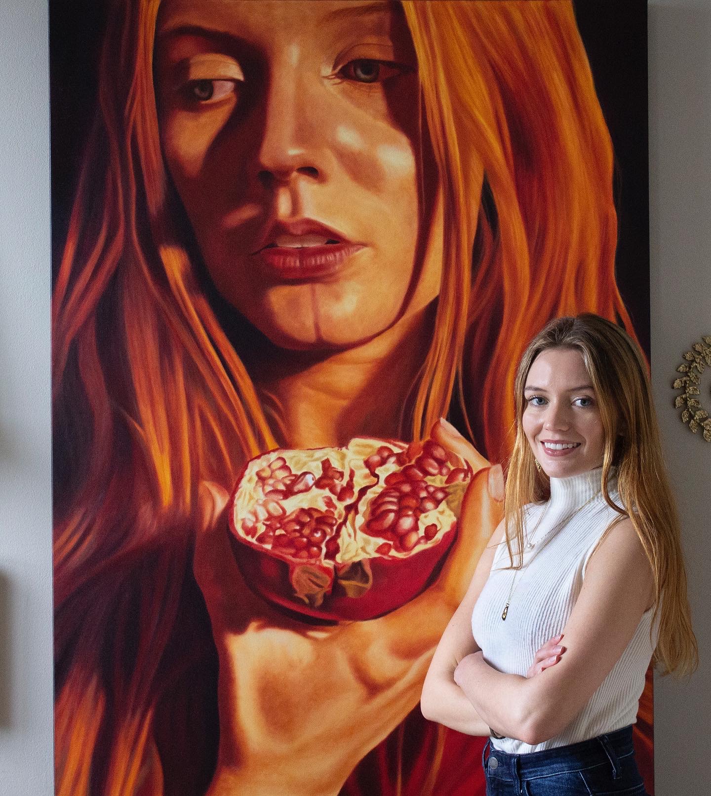 Young woman with long hair smiling, standing with arms crossed in front of a large portrait of a woman with red hair holding a bowl.