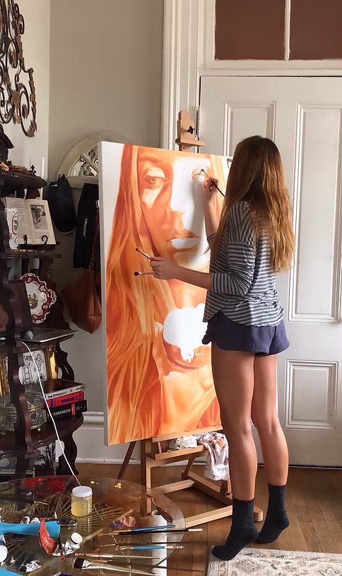 Person with long hair painting a large portrait of a face on an easel in a room.