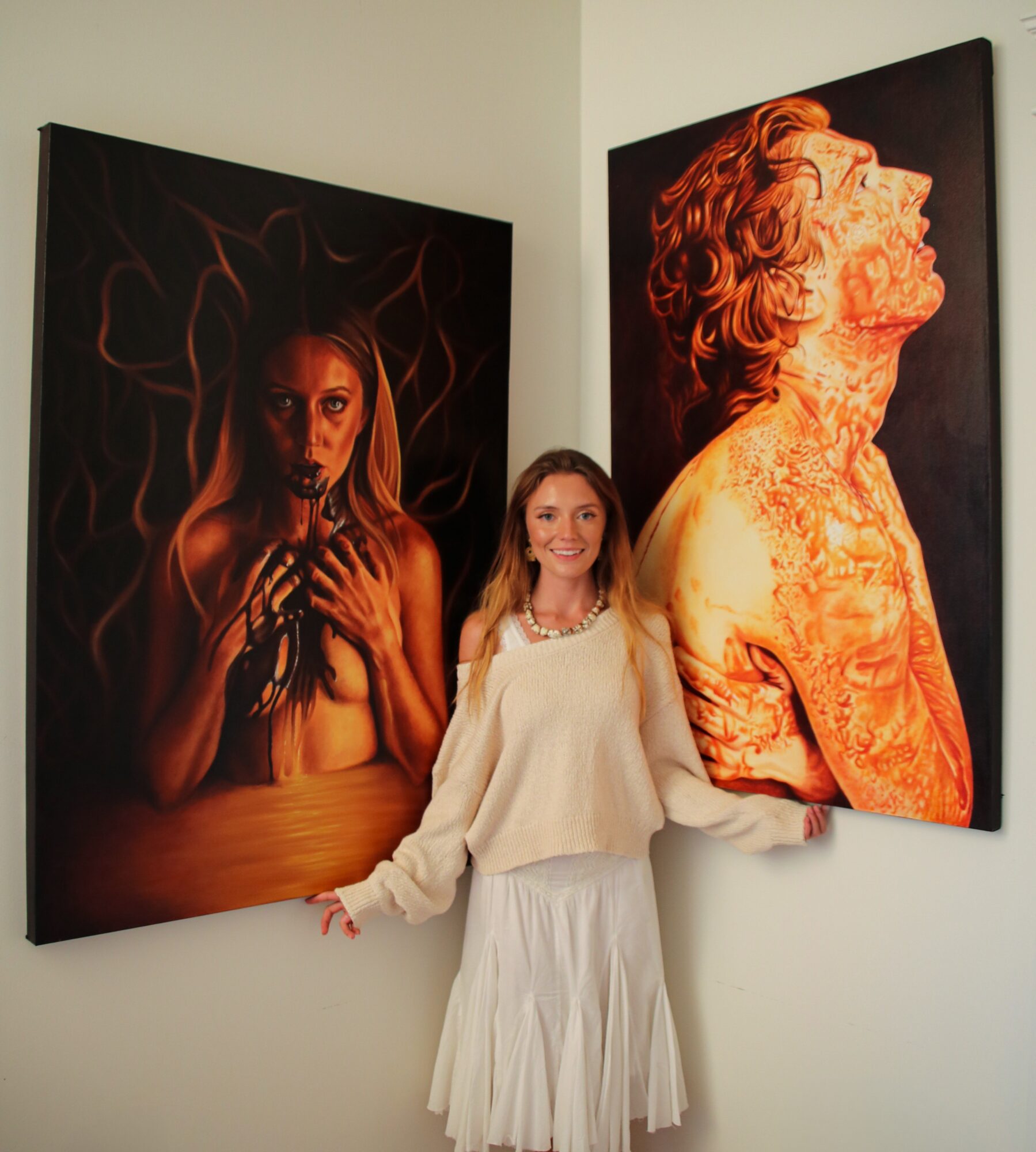 Woman standing between two large paintings on a white wall, smiling, wearing a white top and skirt.
