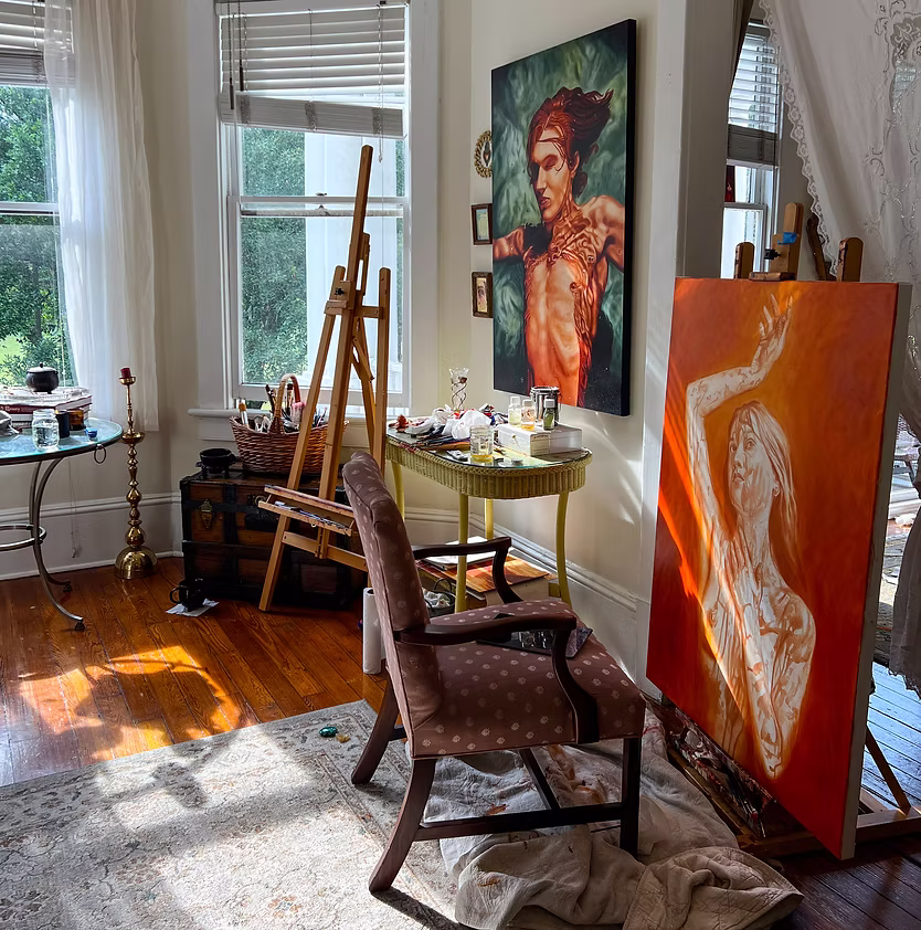 Art studio with paintings, easel, chair, and sunlight streaming through windows.