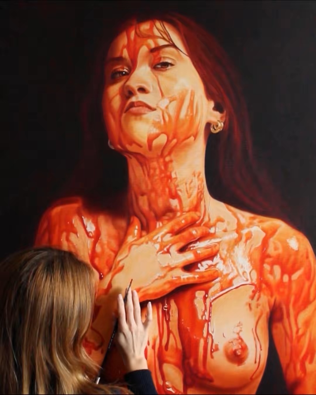 A woman with long hair and a serious expression, covered in red paint or liquid, with a person touching her chest.