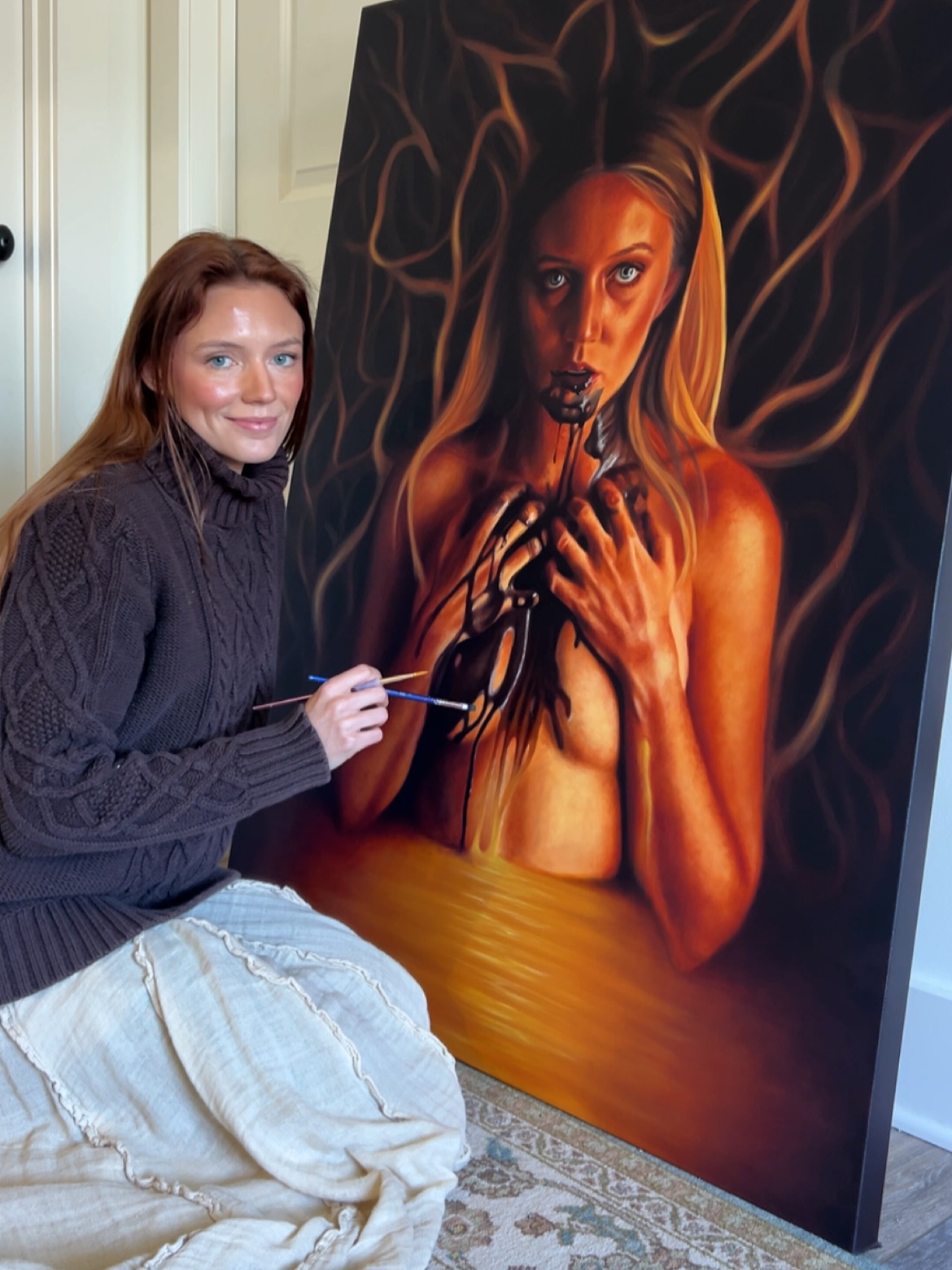 Young woman with brown hair in a dark sweater and light pants, smiling next to a large painting of a woman with fiery hair and hands on chest.